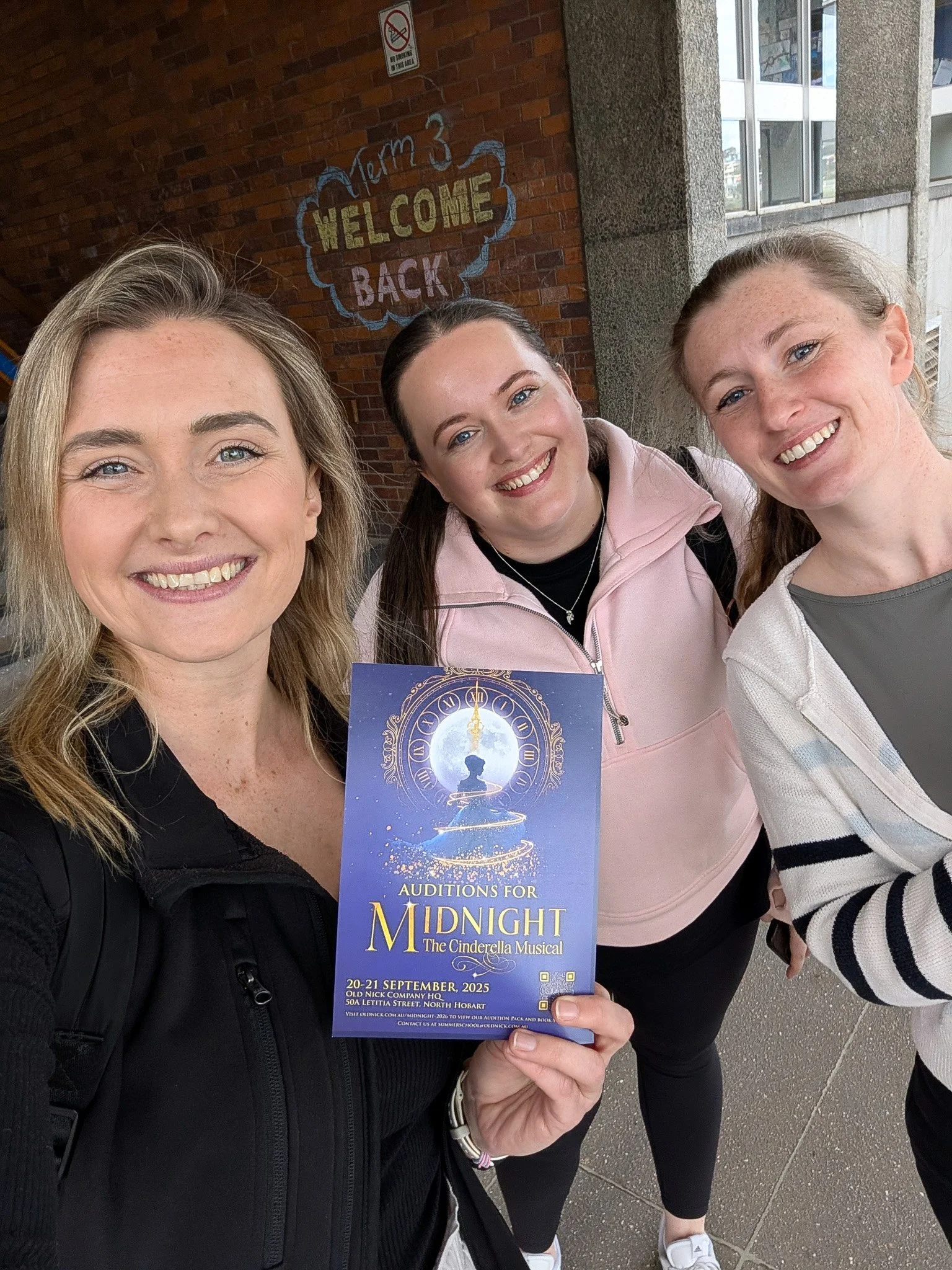 Three smiling women taking a selfie outdoors in front of a brick wall. One woman is holding a blue flyer for a Cinderella musical audition.