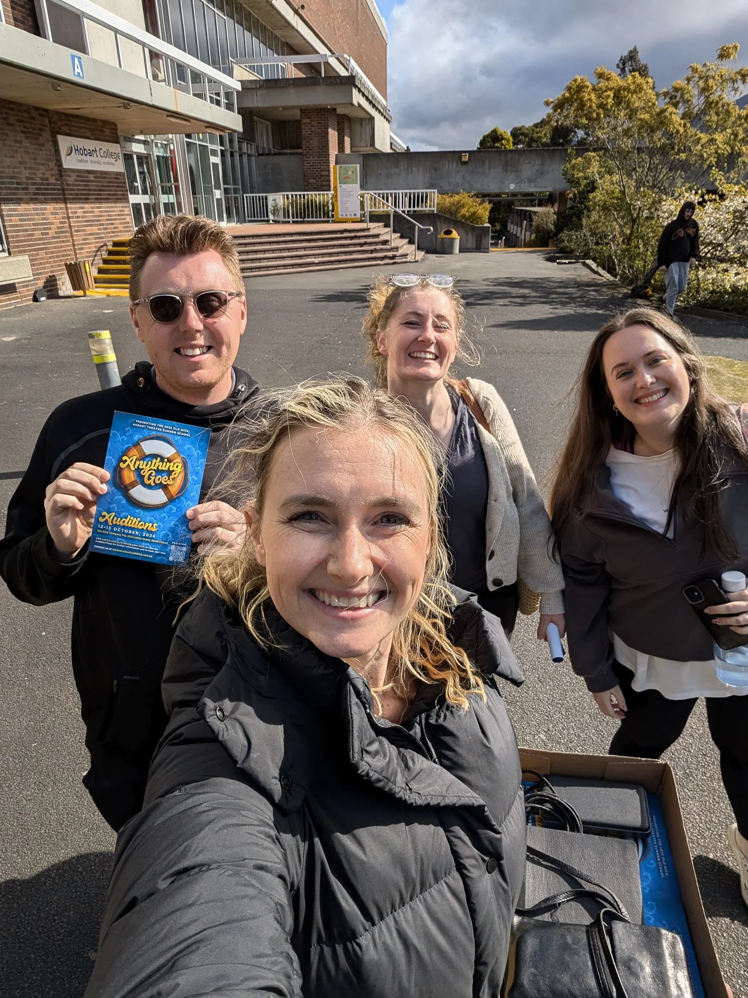 Four people taking a selfie in front of Hobart College with a building in the background, some trees, and a cloudy sky.