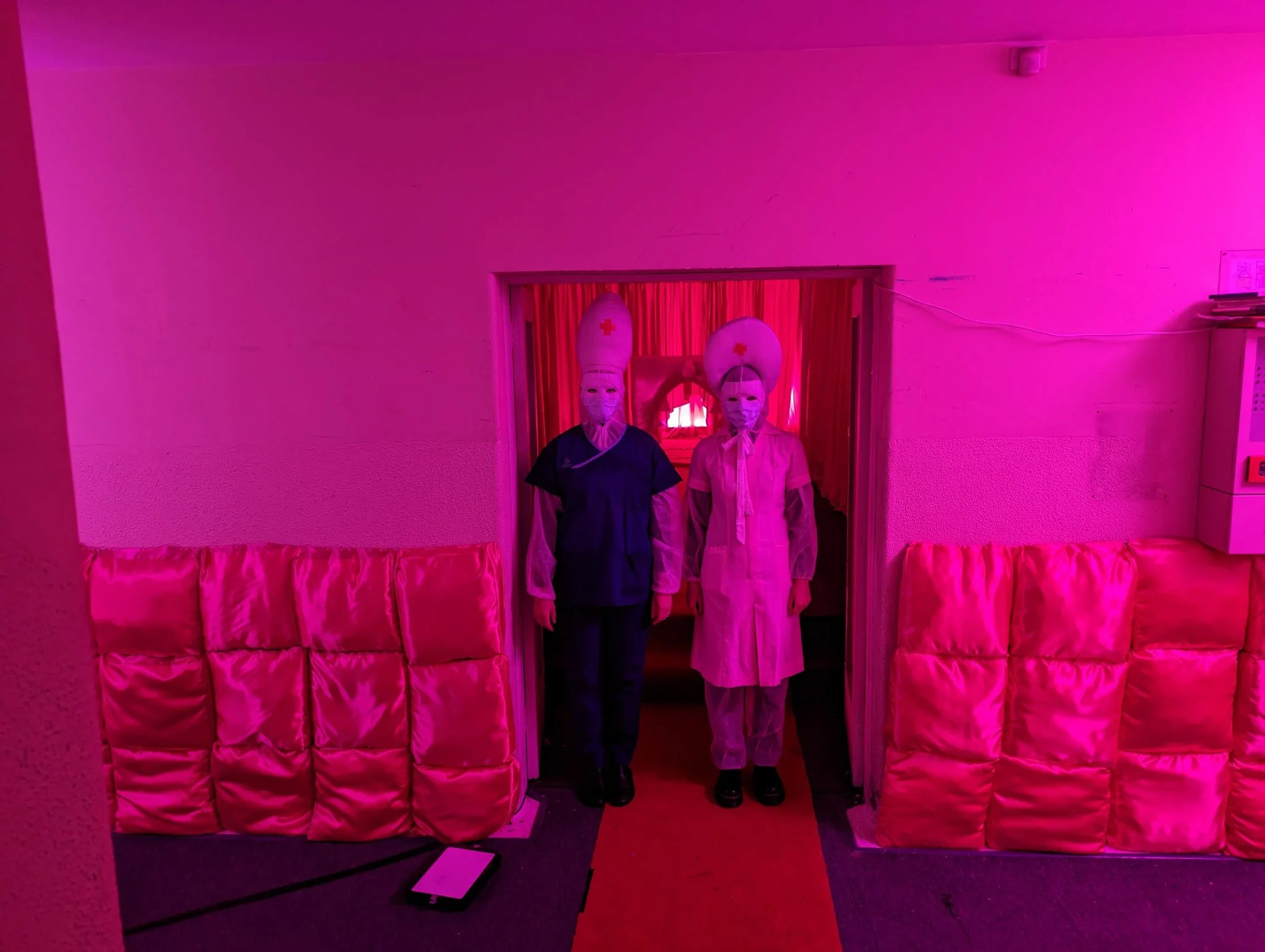 Two people wearing masks and nurse costumes stand in a doorway decorated with pink pillows, with purple lighting.
