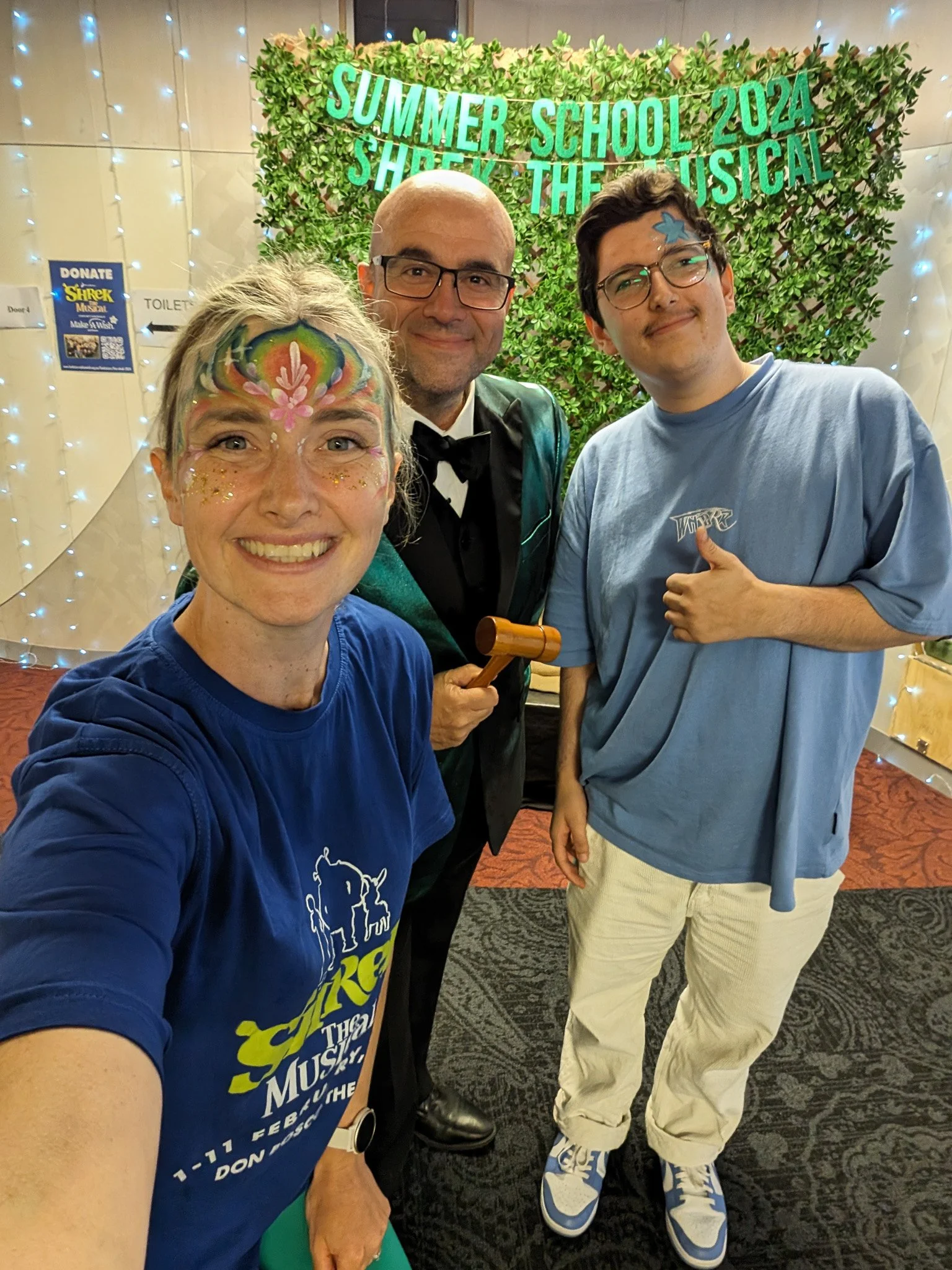 A woman with face paint and glitter smiling, holding a paddle, standing next to two young men in casual clothing. Behind them is a decorated backdrop with green foliage and blue string lights, with a sign indicating a summer school musical theater ev