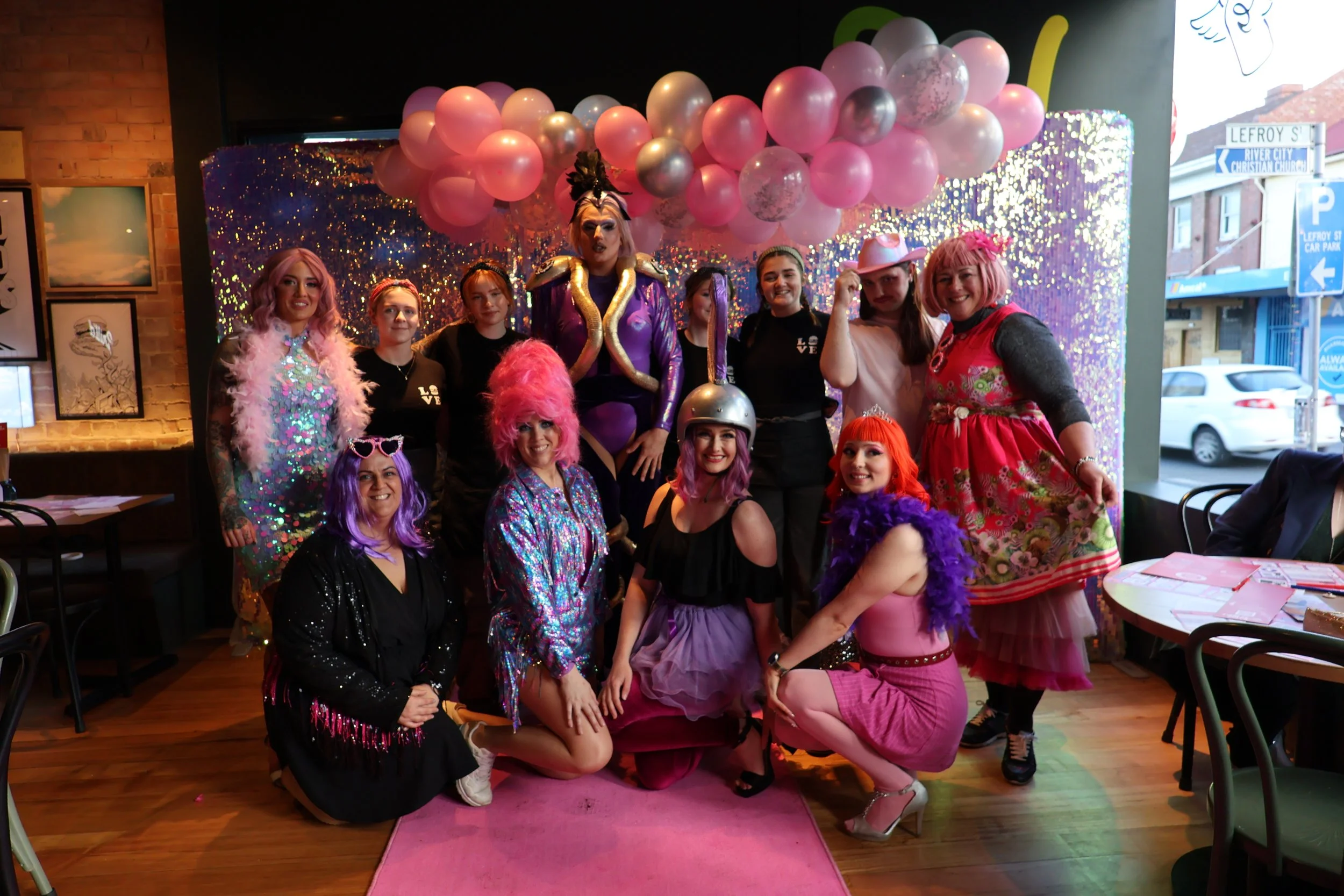 Group of people dressed in colorful costumes, some with pink and purple wigs, posing inside a decorated room with balloons and shiny backdrop.
