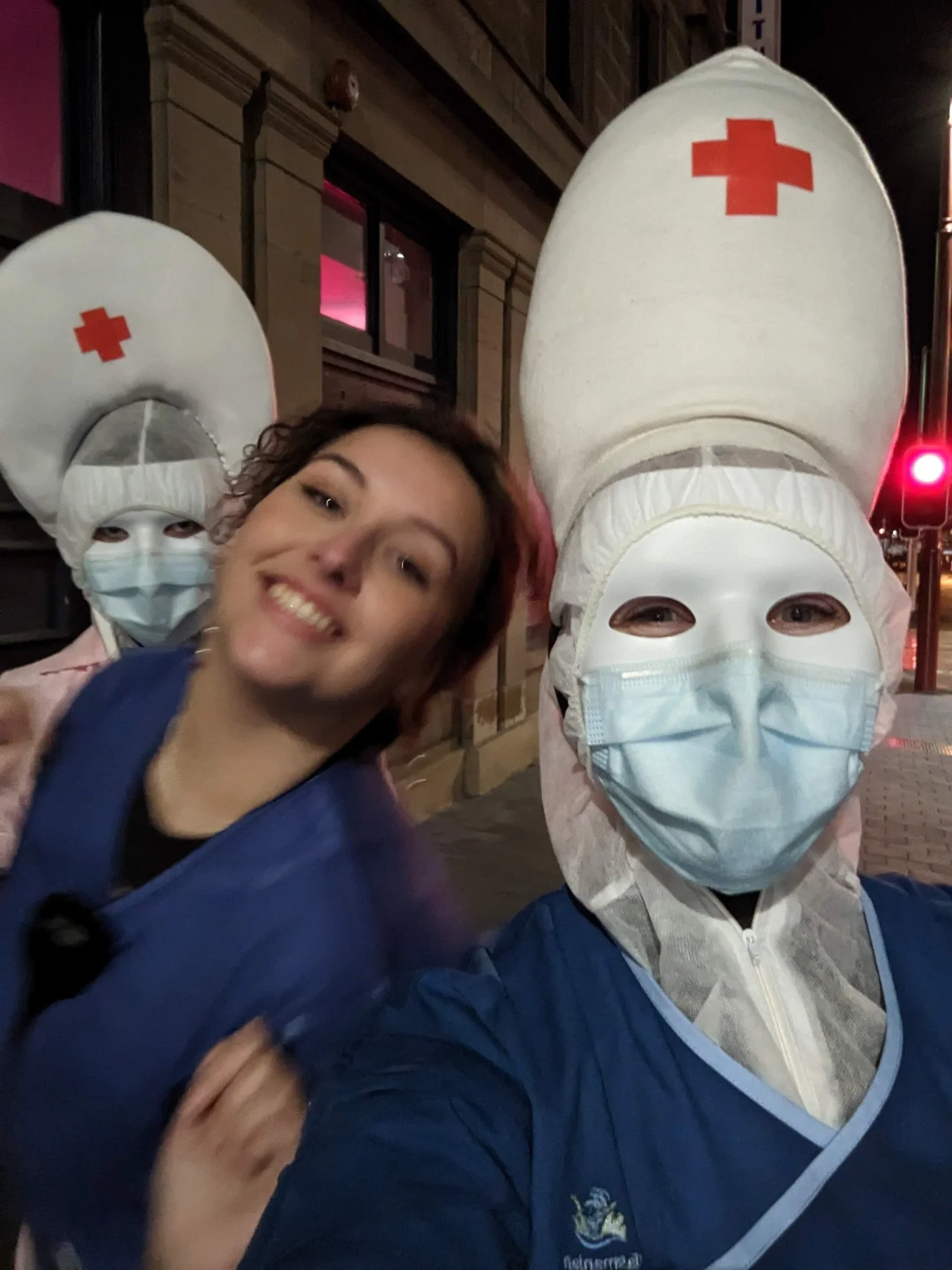 A young woman smiling, wearing a face mask, and taking a selfie with two people dressed in medical uniforms with red cross symbols on their head coverings at night.