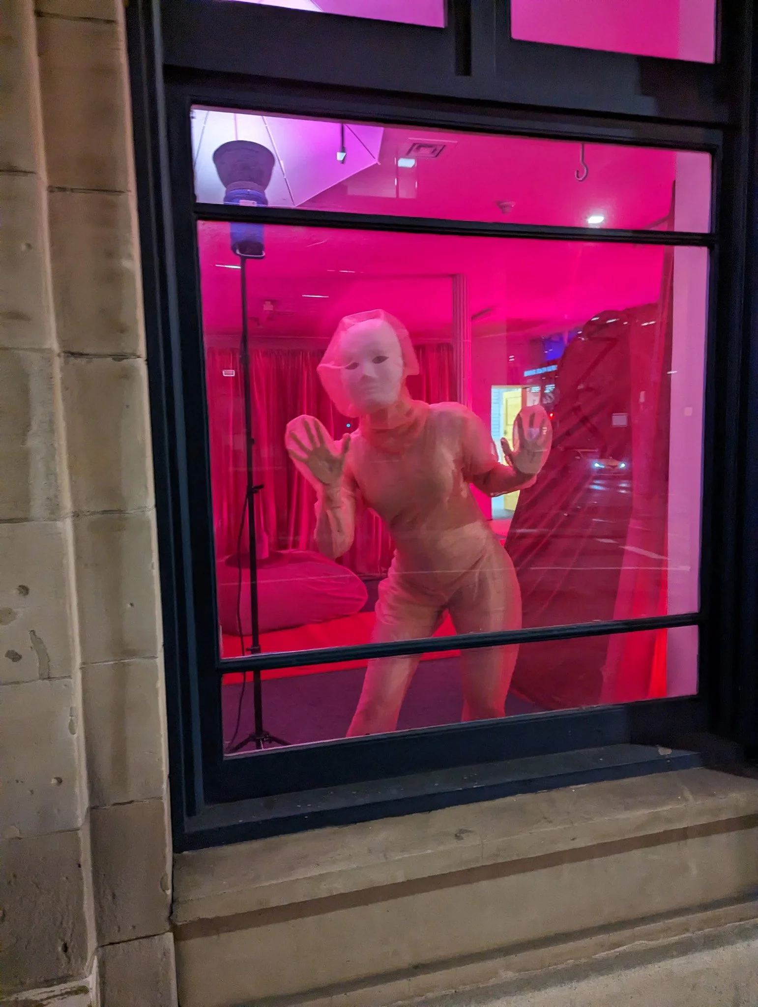 A person dressed in a peach or pink outfit and wearing a white face mask, standing behind a glass window with a pink tint. The person is pressing their hands against the glass and looking through it.