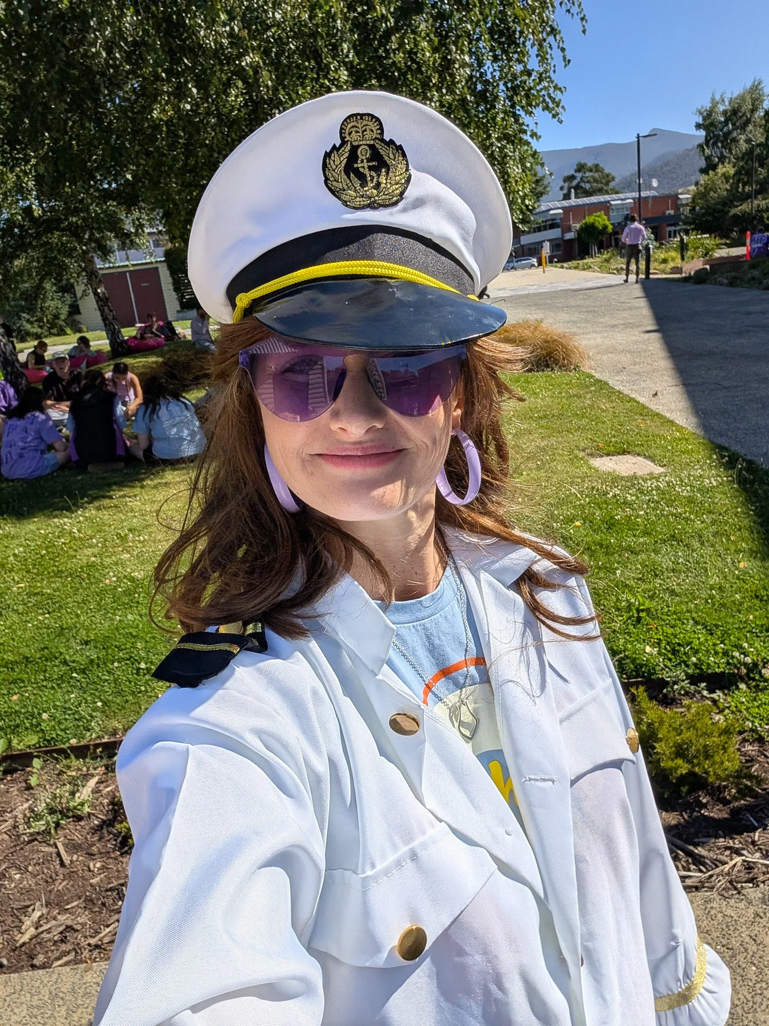 A woman wearing a white Navy captain uniform, including a white hat with a black brim and gold emblem, pink sunglasses, and large hoop earrings, taking a selfie outdoors on a sunny day near a park.