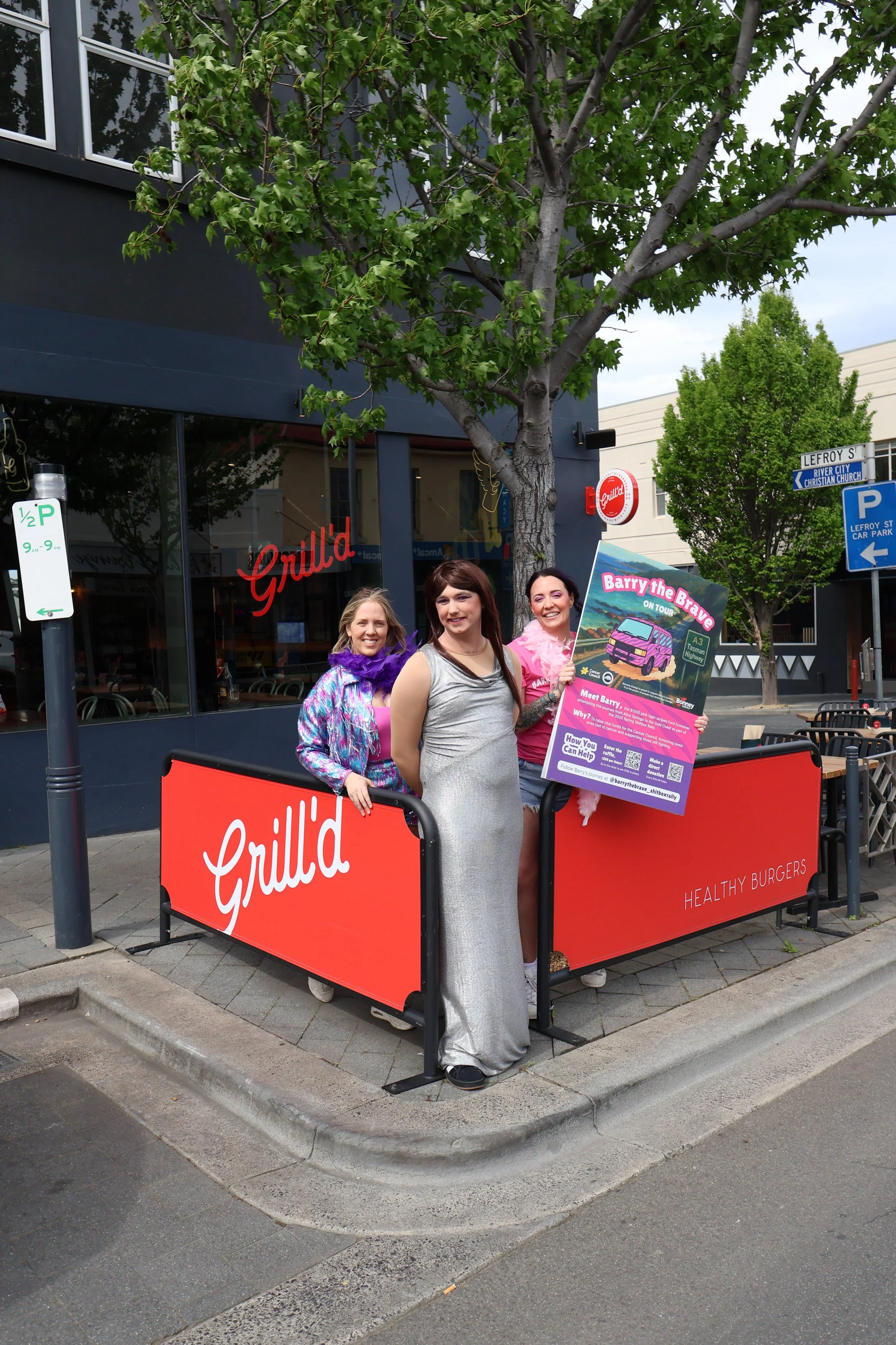 Three women standing behind a red barricade with a sign for a campaign called "Barry the Brave" on a city sidewalk. They are smiling, and one is holding the sign. There are trees, street signs, and a building with a sign for "Grill'd" in the backgrou