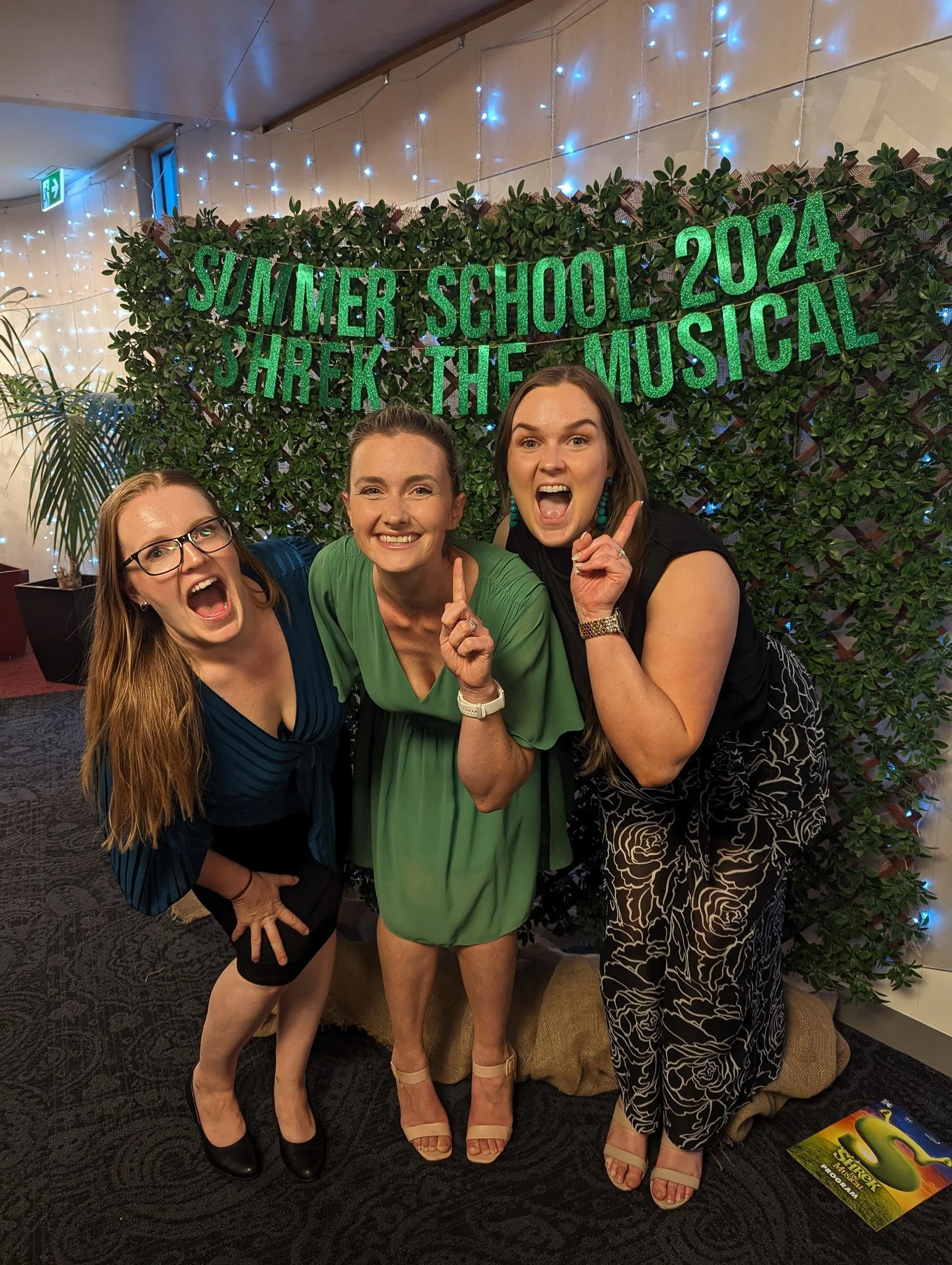 Three women smiling and posing in front of a hedge with a sign that reads 'SUMMER SCHOOL 2024 SHREK THE MUSICAL'. The women are dressed in formal and semi-formal outfits, and the scene appears to be at an indoor event with decorative lighting and pla
