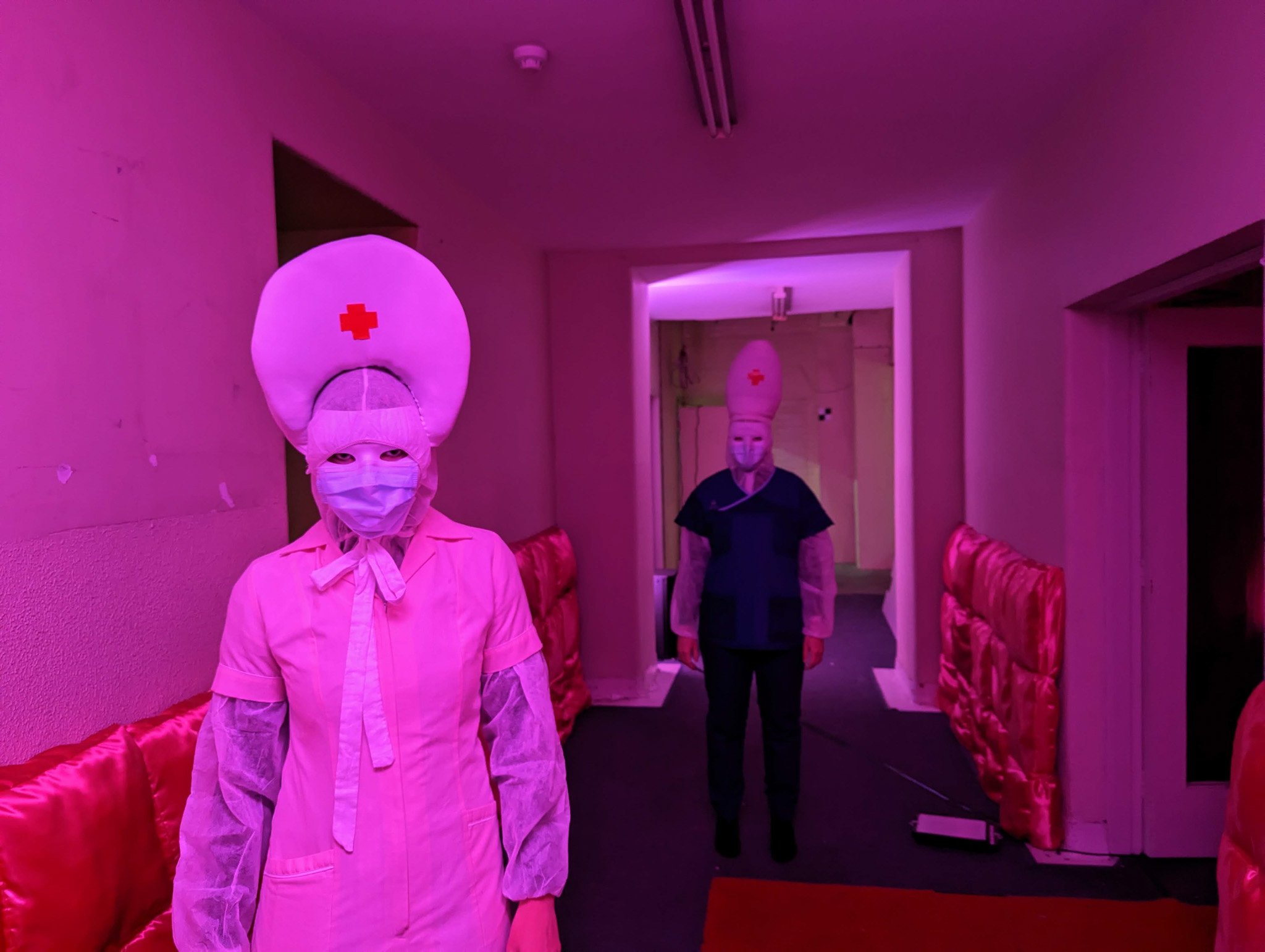Two people wearing masks and costumes resembling medical professionals with red crosses on their hats, in a room with pink and purple lighting, and red cushions along the walls.