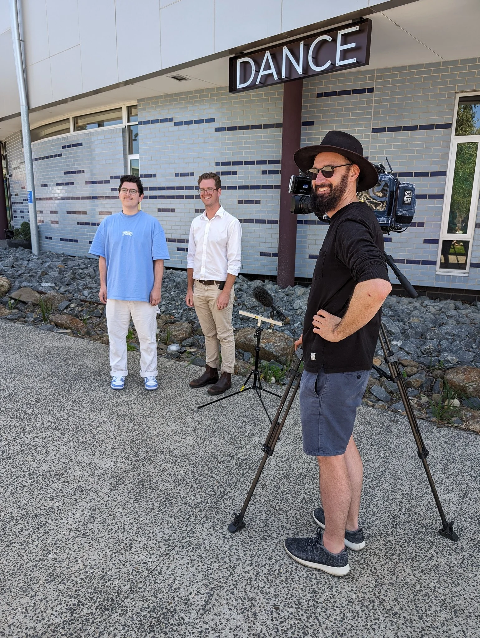 Three men standing outside a building with a sign that says 'DANCE.' One man is operating a video camera on a tripod, and two men are standing nearby, smiling.