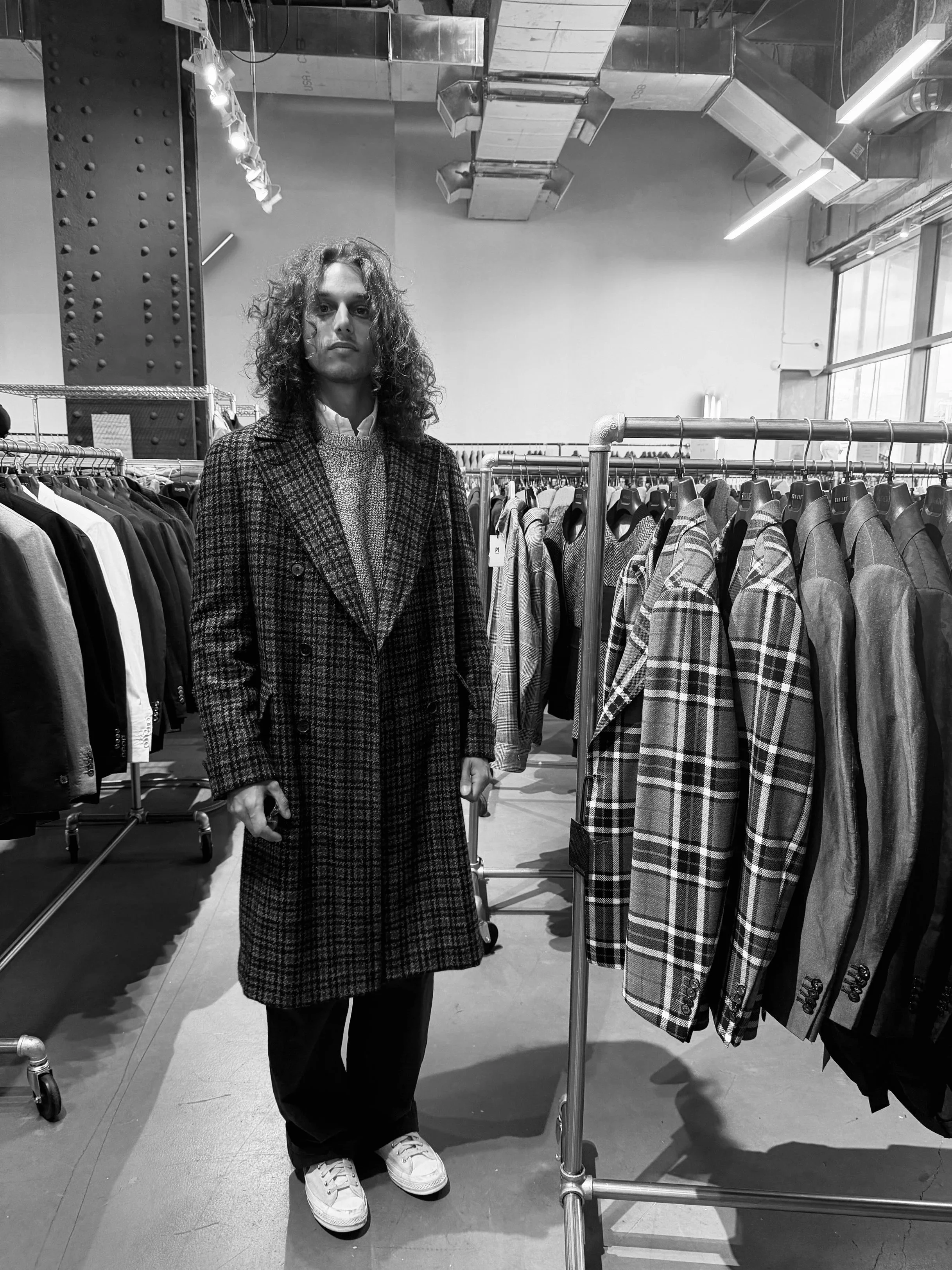 Black and white photo of a person with curly hair standing in a clothing store between racks of jackets and coats.