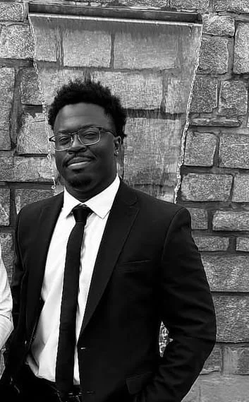 A young man with glasses and curly hair in a suit, standing outdoors in front of a stone wall.