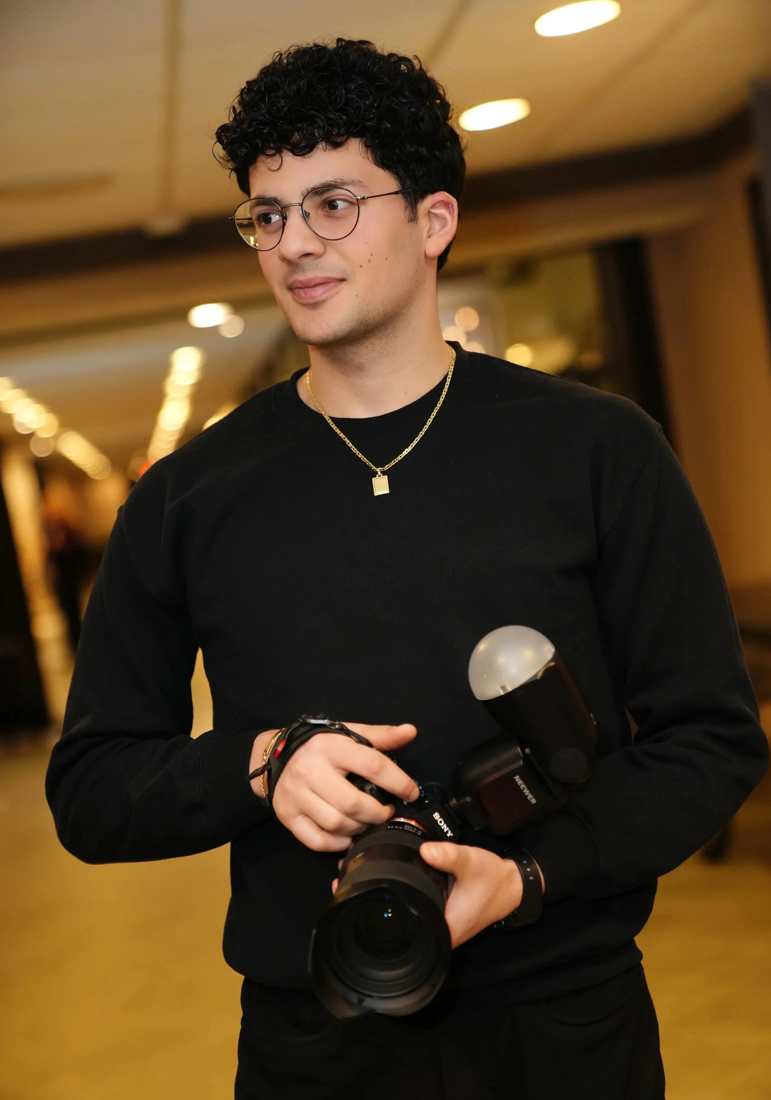 Jeune homme avec lunettes, cheveux bouclés, tenant un appareil photo en intérieur, portant un bracelet et une chaîne