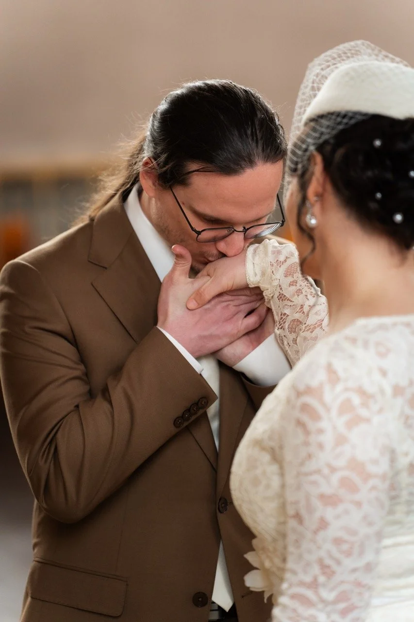 Un couple de mariés s'embrassant lors de leur mariage, la femme portant une robe en dentelle et un voile, tenant un bouquet de fleurs, en pleine journée devant un bâtiment en brique.