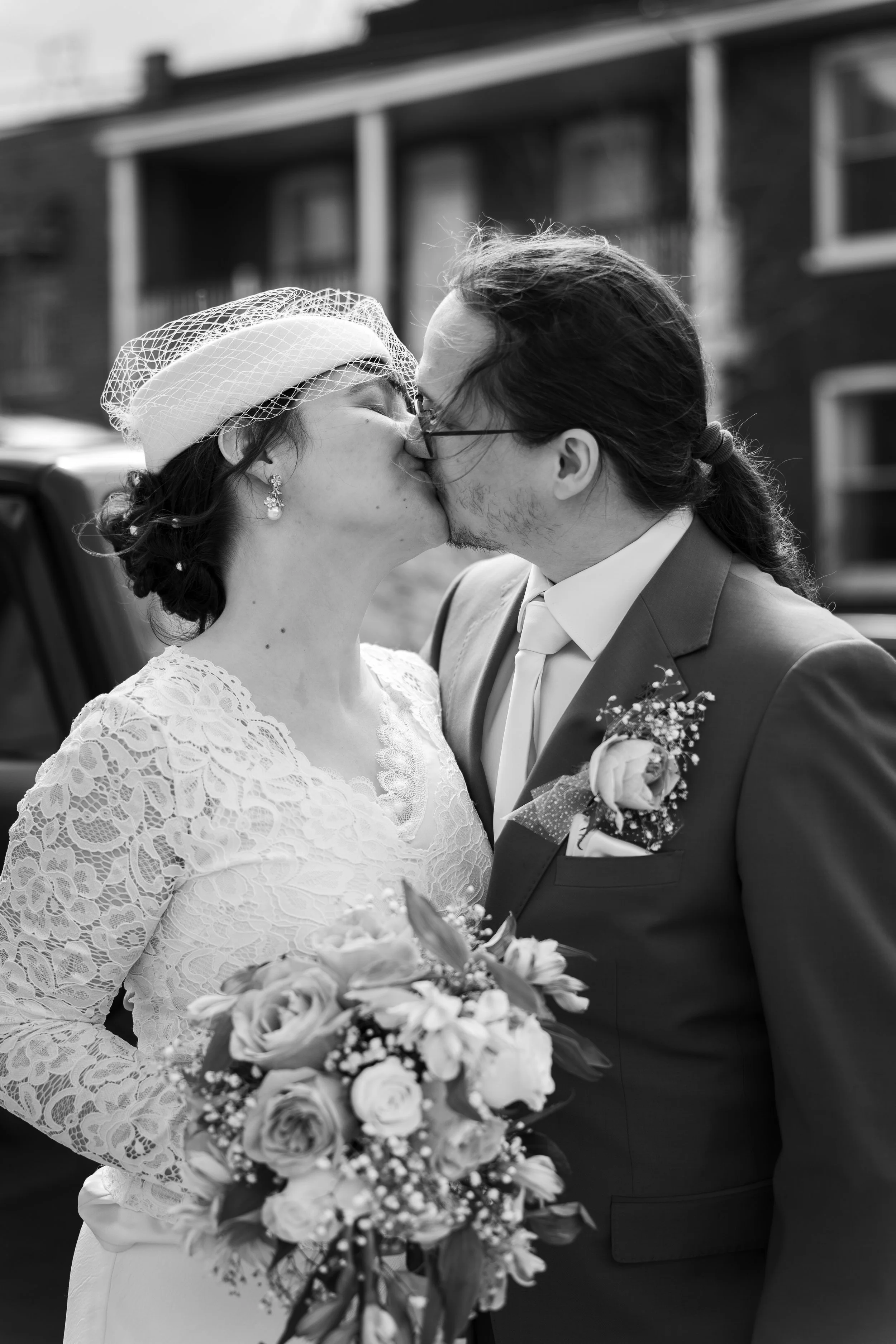 Un couple de mariés s'embrassant lors de leur mariage, la femme portant une robe en dentelle et un voile, tenant un bouquet de fleurs, en pleine journée devant un bâtiment en brique.