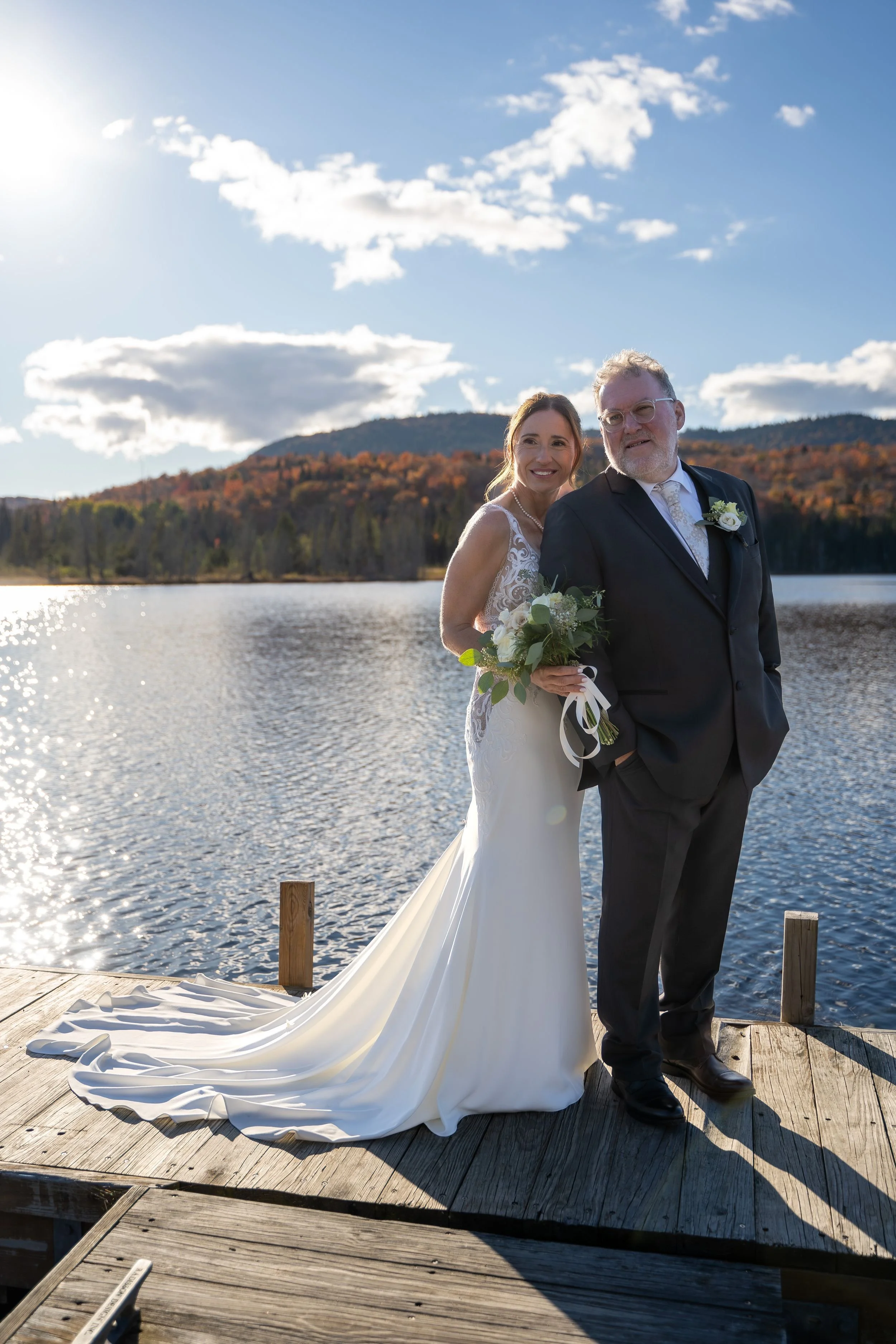 Un couple en tenue de mariage sur un pont en bois au bord d'un lac, avec une montagne en arrière-plan et un ciel ensoleillé avec quelques nuages.