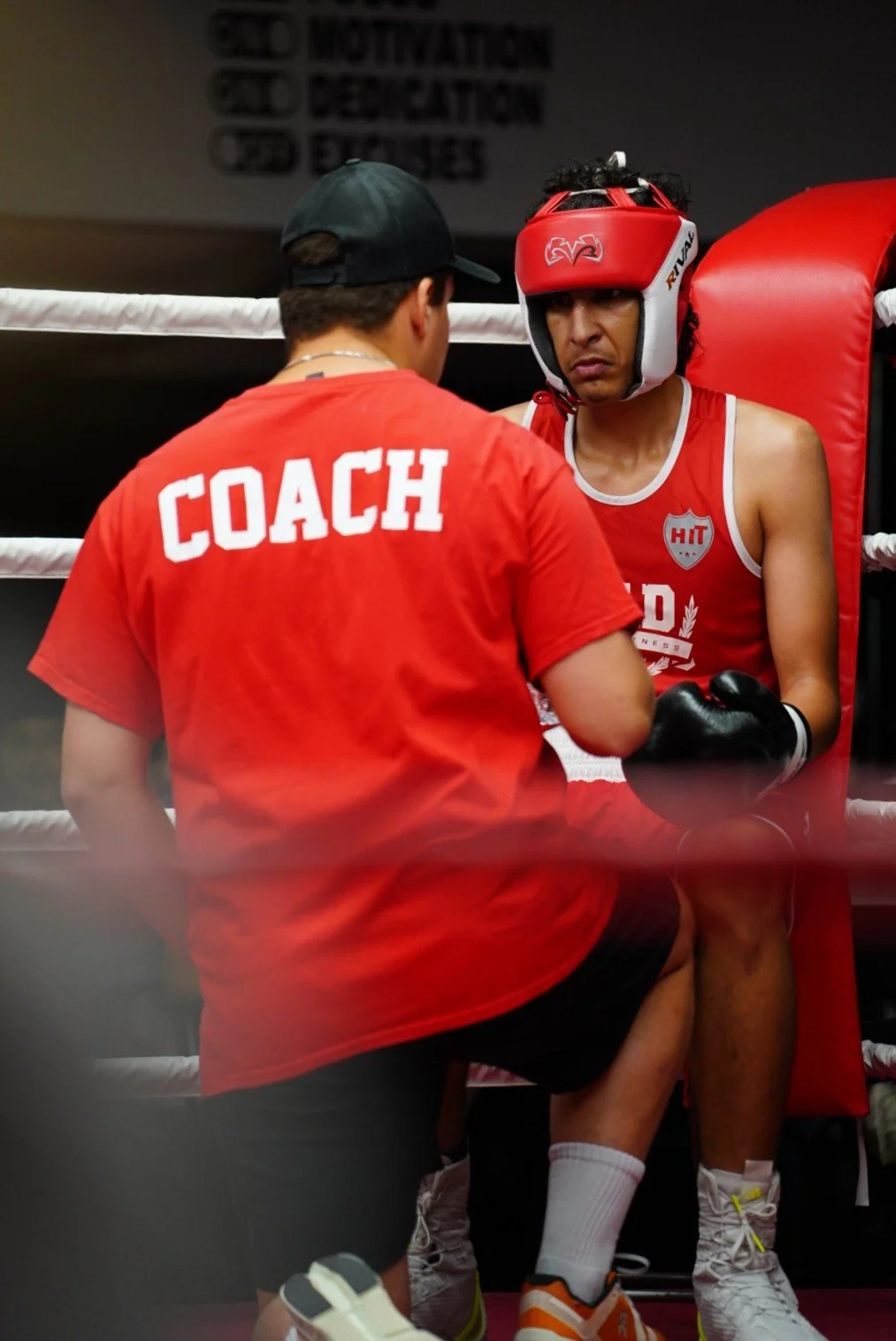 Un coach parle à un boxeur assis dans un coin du ring, portant un casque de boxe rouge, vêtements de boxing rouges et gants noirs.