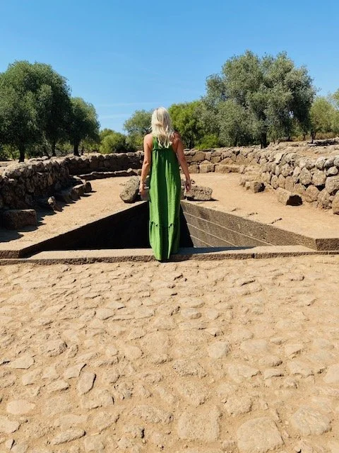 Una donna con abito verde chiaro che cammina su un sito archeologico con abbeveratoio di pietra e alberi sullo sfondo sotto un cielo azzurro