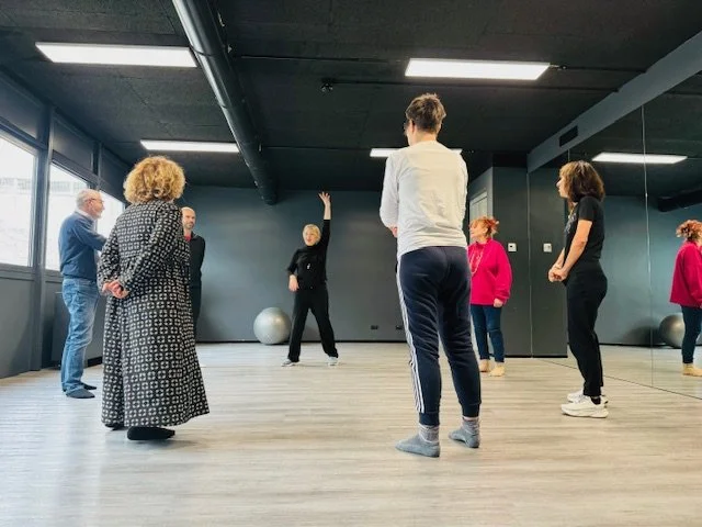 Gruppo di persone in una sala di danza, alcune osservano, altre si preparano a ballare, con un grande specchio sulla parete.