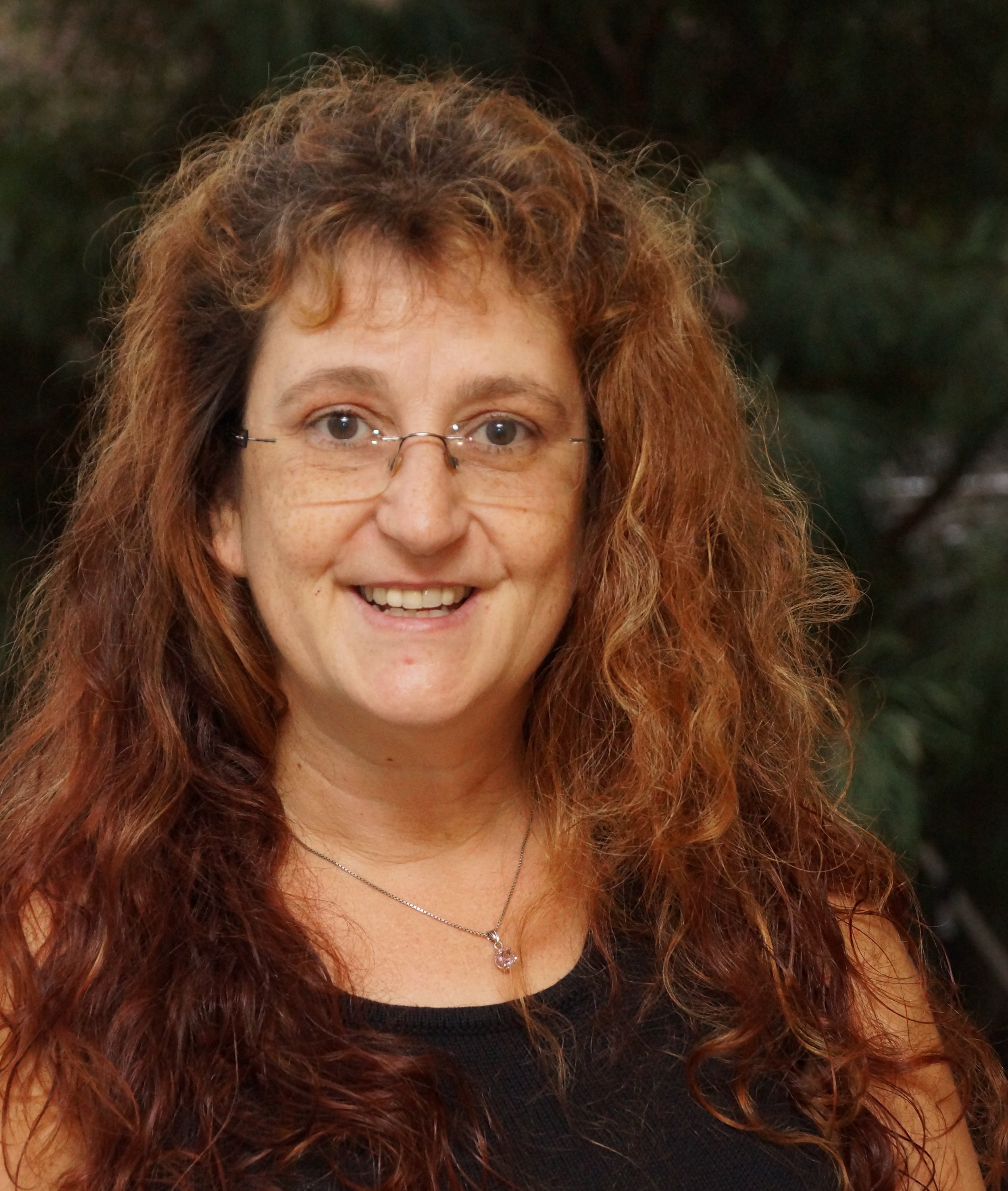 Close-up of a woman with curly red hair, glasses, wearing a black top and a necklace with a pink pendant, smiling.