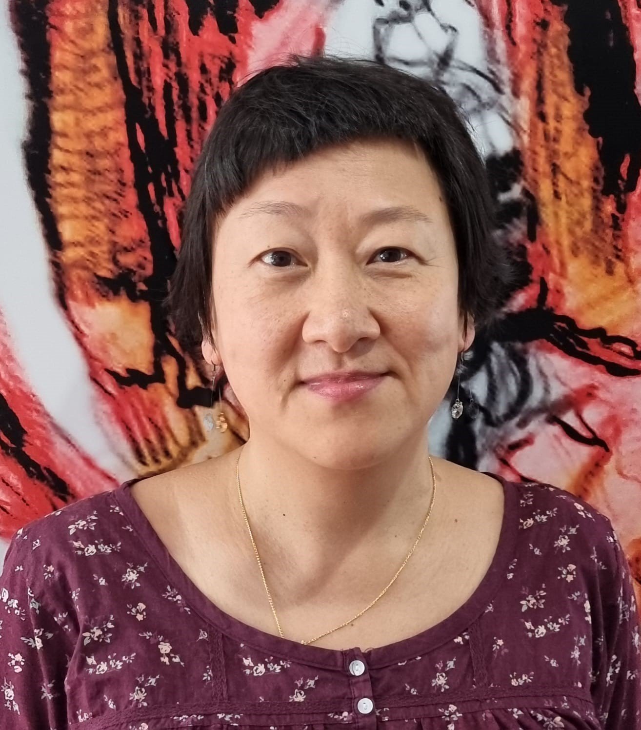 A woman with short wavy dark hair wearing a burgundy floral blouse, earrings, and a gold necklace, standing in front of a colorful textile with tiger stripes.