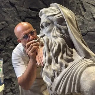 A man with glasses sculpting or examining a stone bust of an ancient-looking bearded man with a head covering, set against a rocky background.