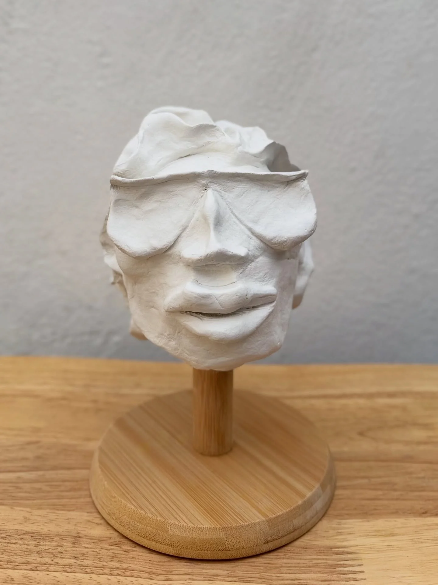 White sculpted face with closed eyes, lips slightly parted, mounted on a wooden stand.