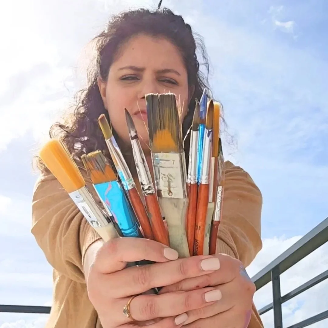 A woman holding a bunch of paintbrushes towards the camera, outdoors with a blue sky and clouds in the background.
