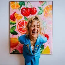 A woman with blonde hair smiling and laughing in front of a colorful fruit-themed painting, wearing a bright blue outfit.