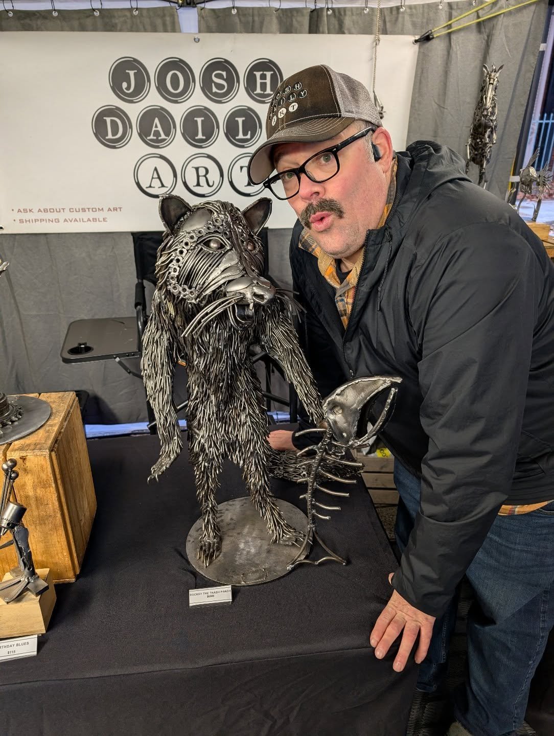 Man wearing glasses, a cap, and a black jacket posing with a metallic sculpture of a lion holding a cup, displayed on a table at an art booth.