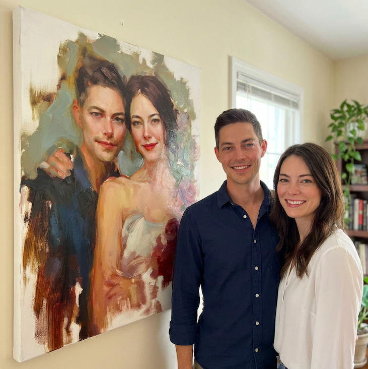 A smiling man and woman stand together in front of a large portrait painting of themselves, hanging on a beige wall in a room with a window and a bookshelf with plants.