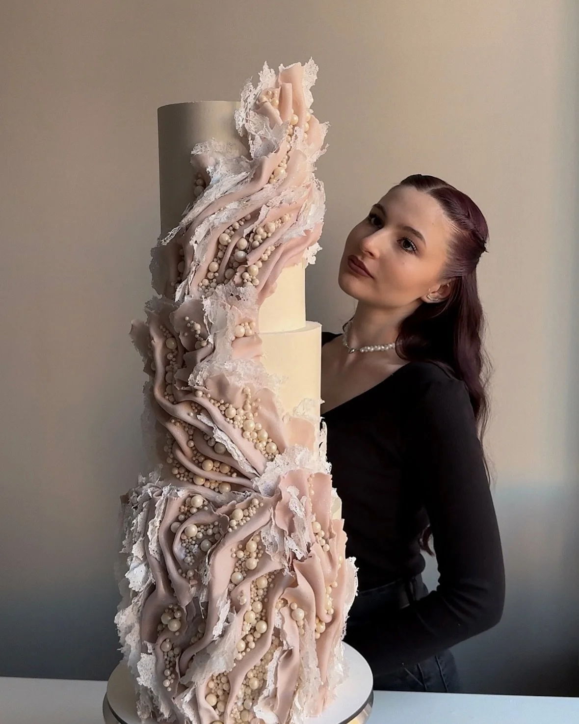 A woman with long, wavy dark red hair in a black top and pearl necklace stands behind a tall, elaborate cake with a neutral plain background.