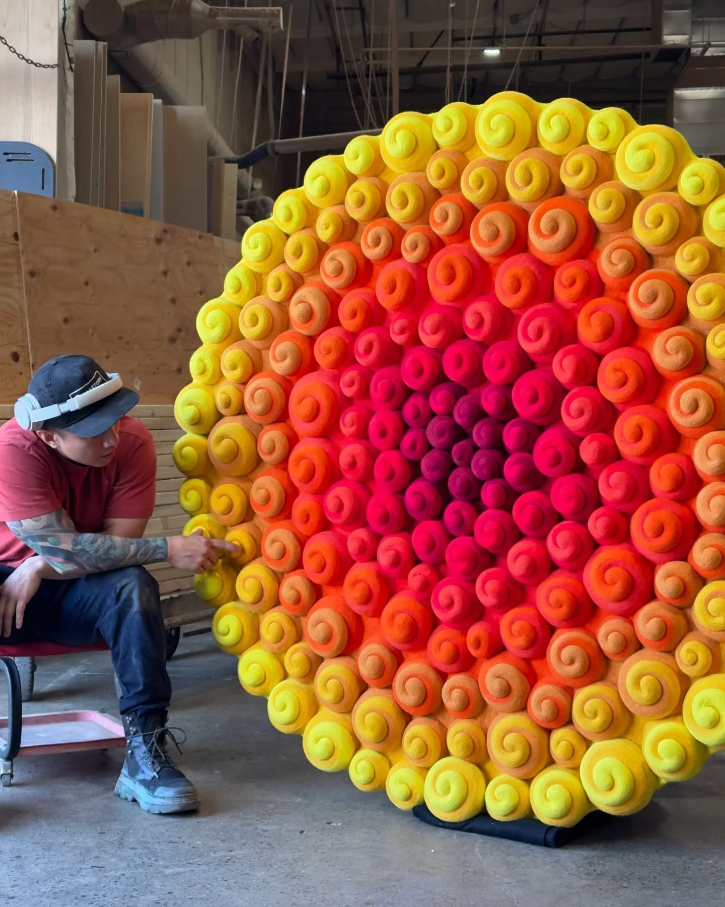 A person working on a large, colorful, spiral-shaped art installation made of rolled fabric or foam in a workshop.