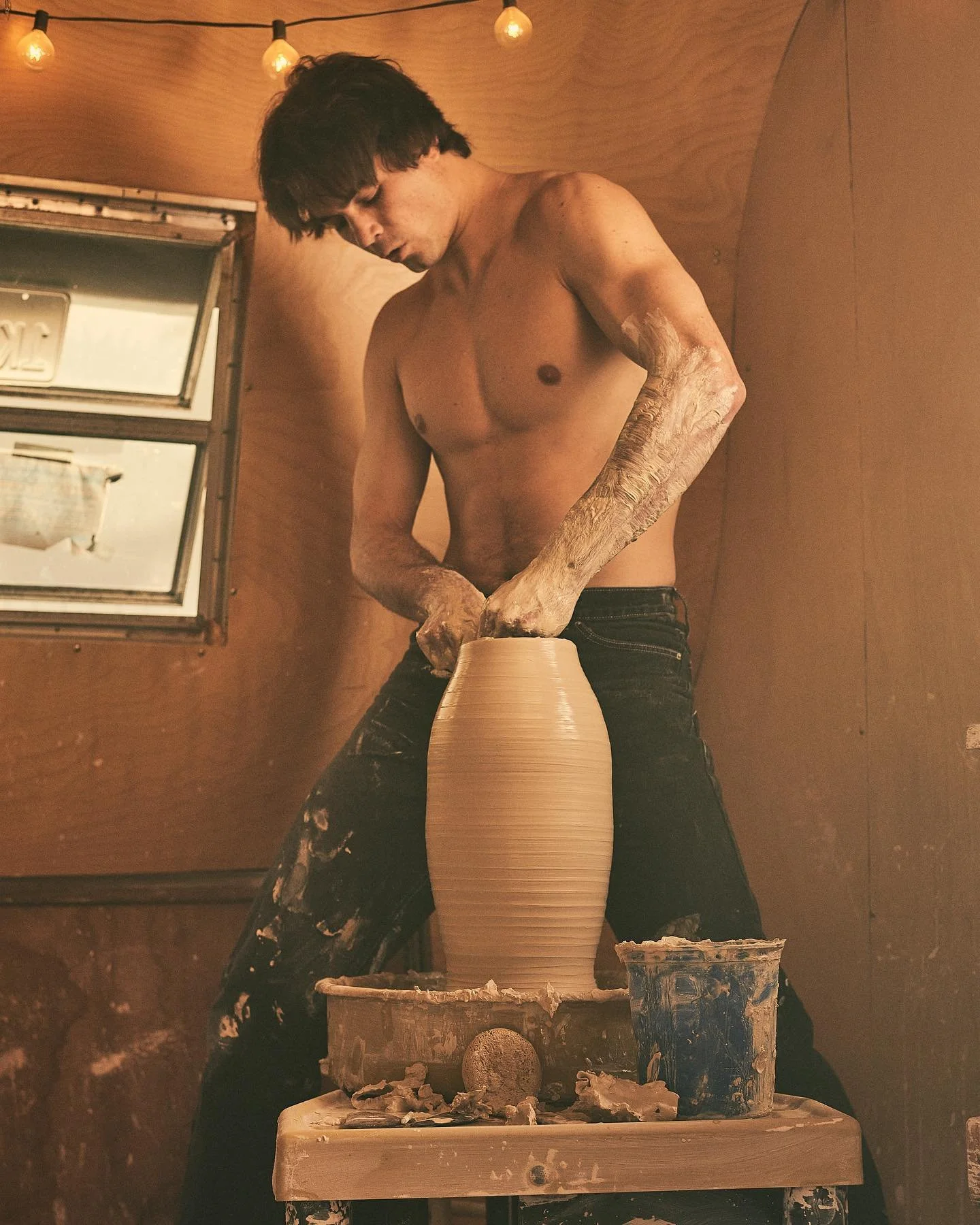 A shirtless man with dark hair and tattoos on his arms is shaping a tall ceramic vase on a pottery wheel in a warm, wood-paneled room with string lights.