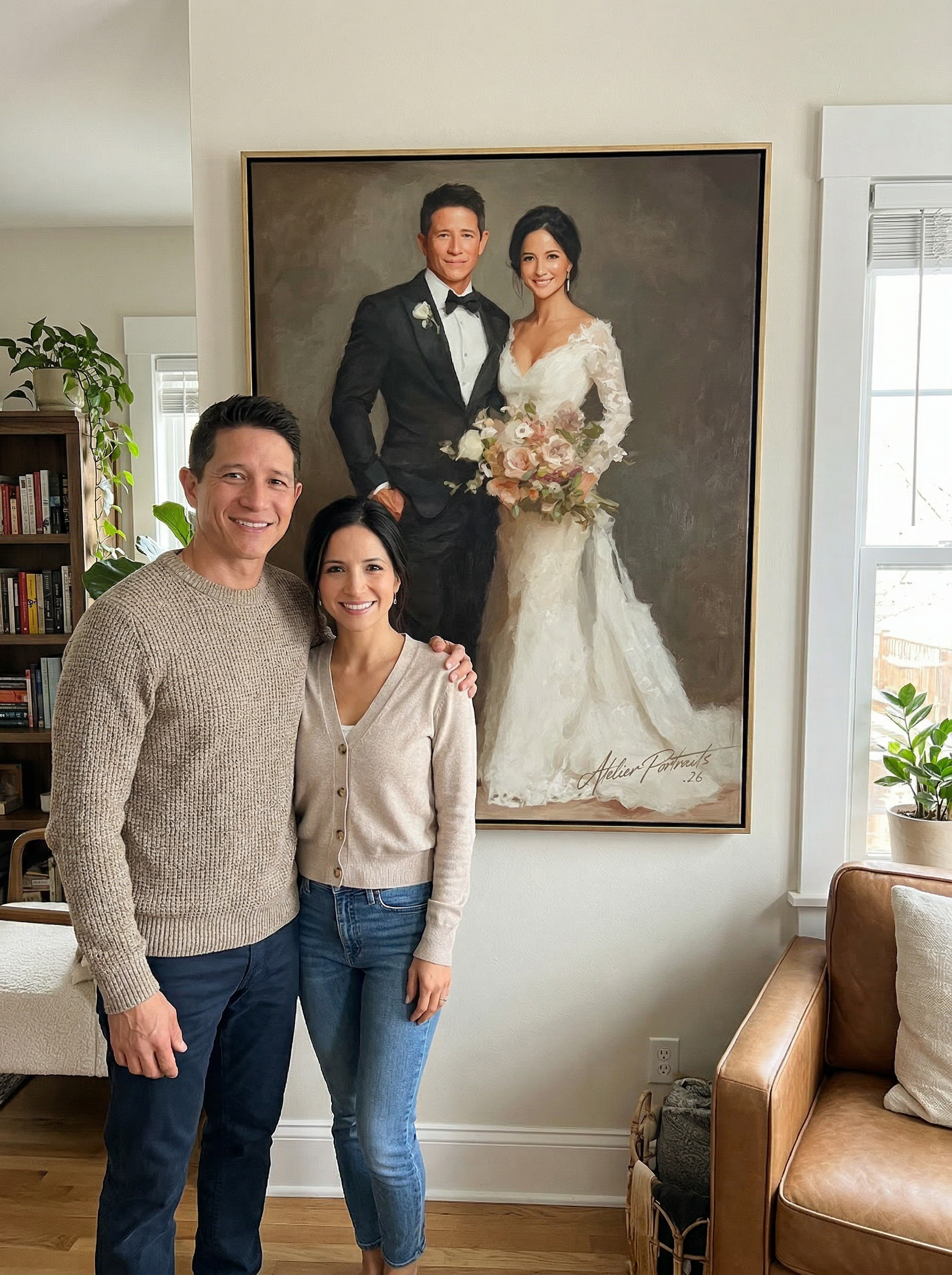 A man and woman smiling and standing together in front of a large wedding portrait of a bride and groom. The portrait shows the couple in wedding attire, with the bride holding a bouquet. The room has a window with natural light, a bookshelf, and a leather couch.