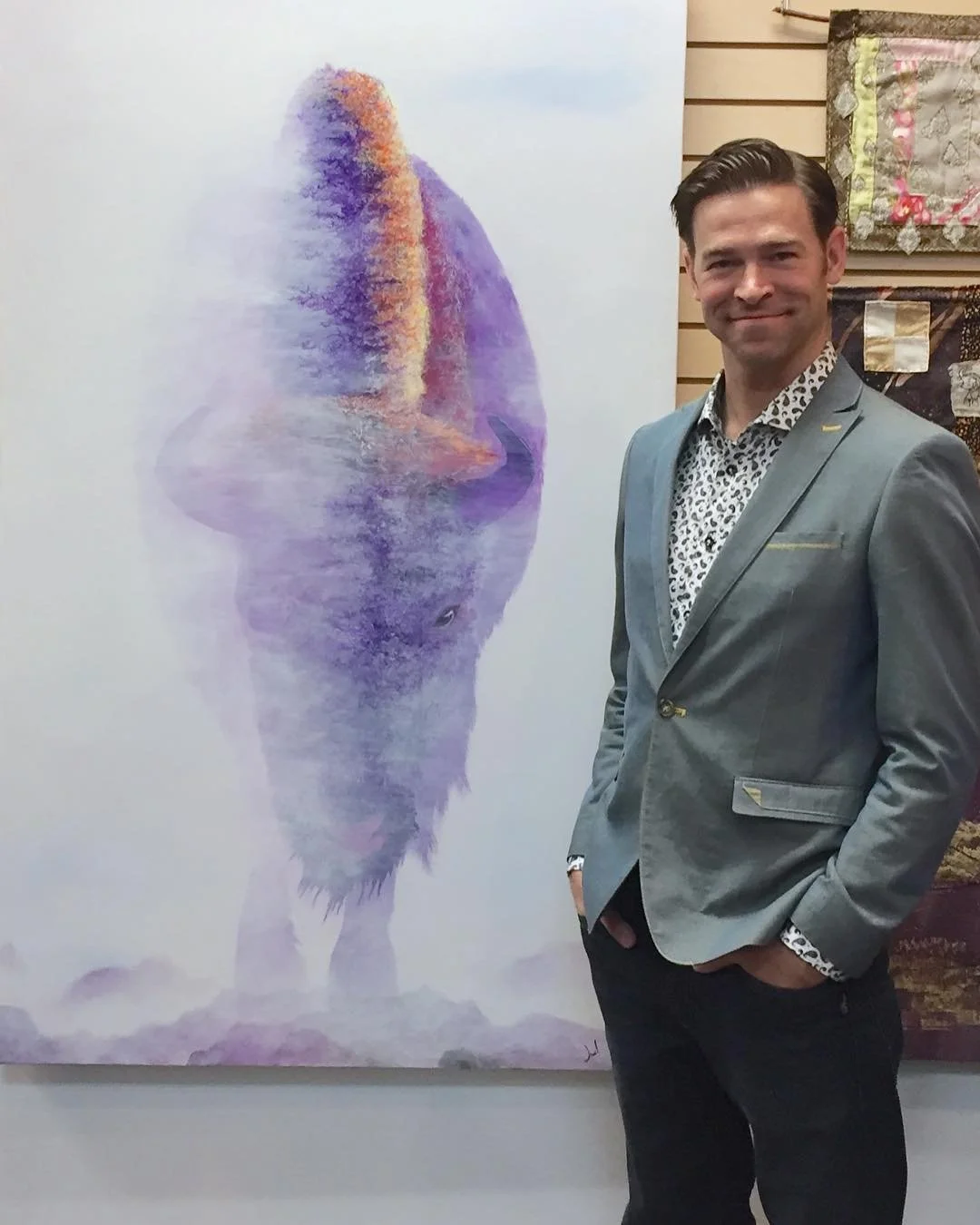 A man in a light grey suit jacket and patterned shirt stands next to a large watercolor painting of a wolf, with the man posing and smiling.