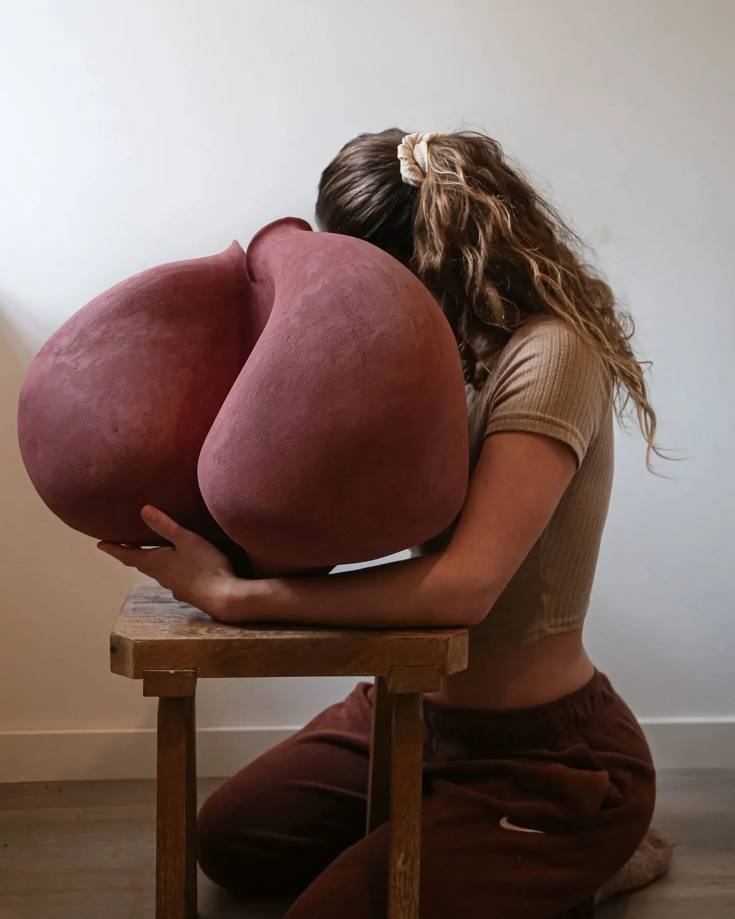 A woman kneeling on the floor holding a large, heart-shaped sculpture made of reddish-brown material.