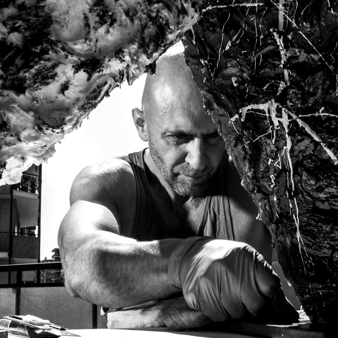 Man with a shaved head and beard, wearing a sleeveless shirt and gloves, slicing large piece of raw meat on a table outdoors.