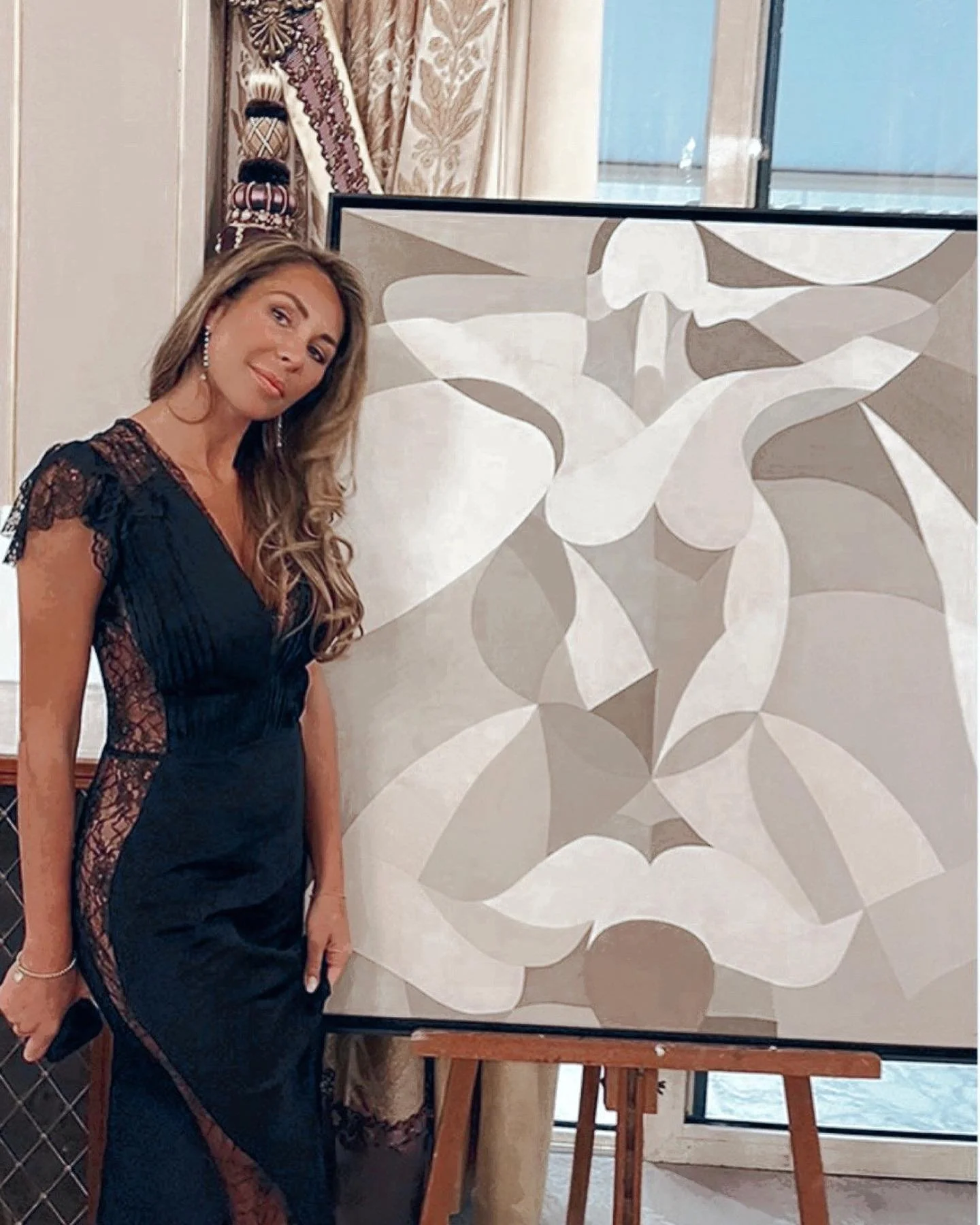 A woman with long wavy hair in a black dress with lace details, standing next to an abstract painting on an easel. The background shows elegant drapes and a window.