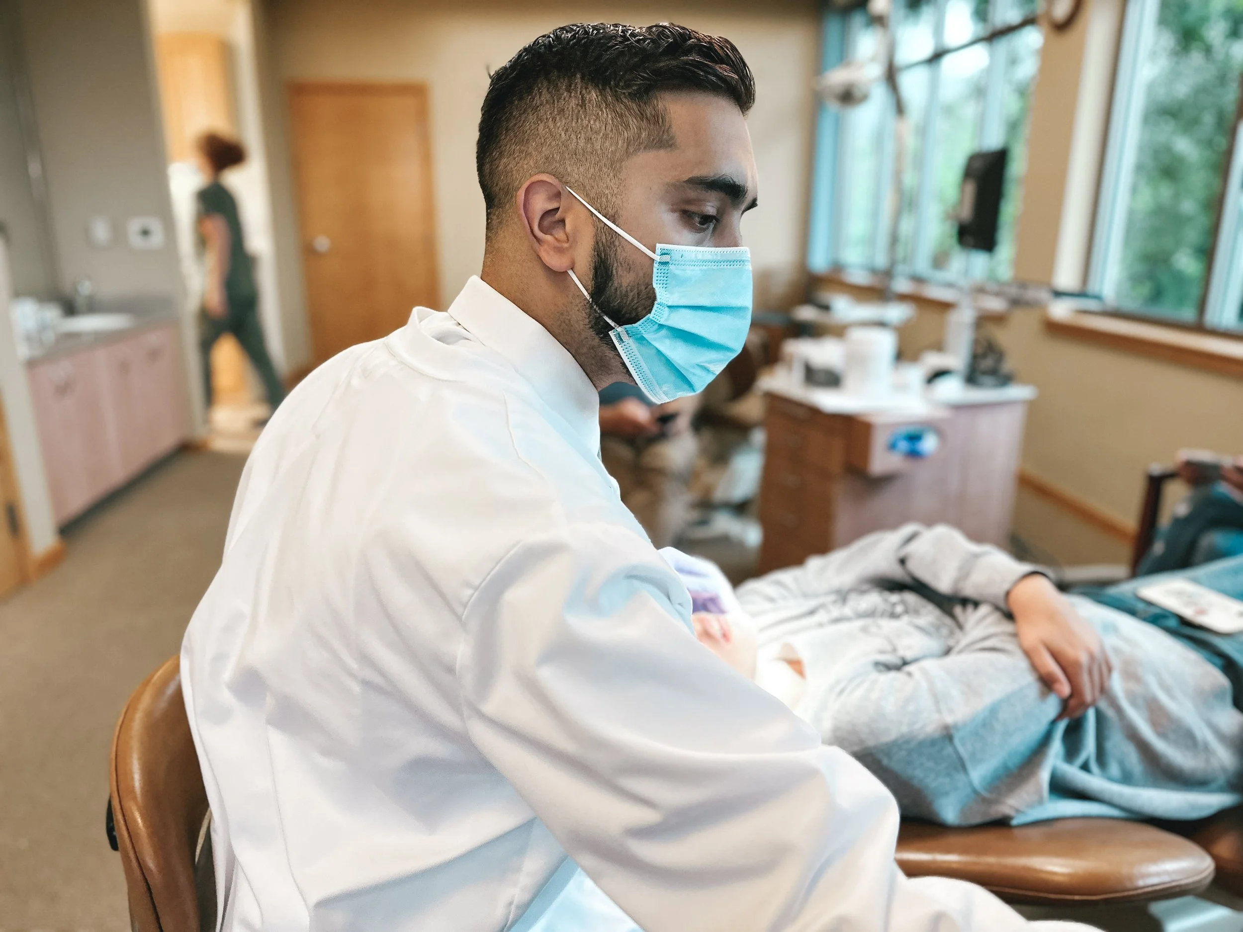 A healthcare professional wearing a surgical mask and white coat attending to a patient in a medical facility.