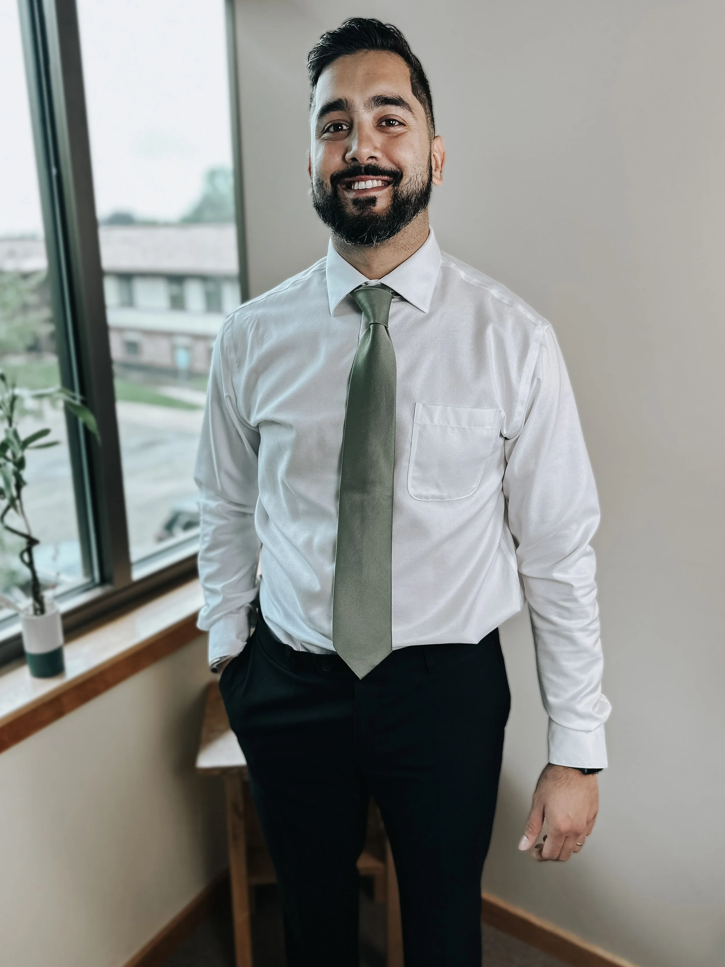 A man with dark hair and a beard dressed in a white shirt and black pants standing in an office with a window showing buildings outside.