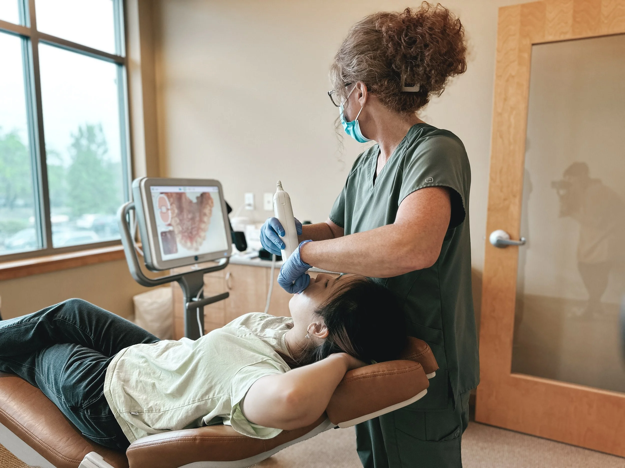 A healthcare professional performing a dental procedure on a patient lying on a dental chair in a clinic. The professional is wearing scrubs, gloves, a face mask, and glasses, and is using a dental instrument. The patient has their mouth open. There is a computer screen nearby showing dental images and a window with a view outside.