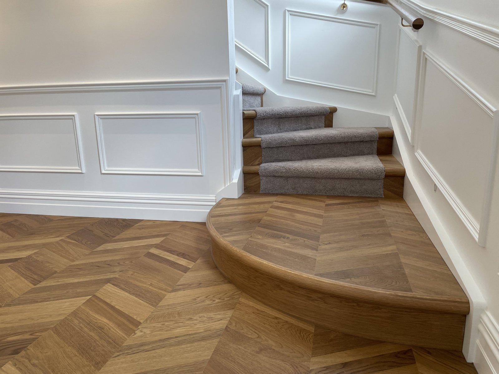 Interior view of a staircase with beige carpeted steps, white paneled walls with molding, and wood flooring with a chevron pattern. There is a curved wooden railing and a partial view of the landing.