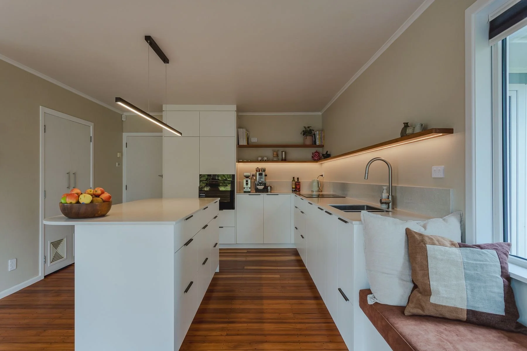 Modern kitchen with white cabinets, wooden shelves, a countertop with a coffee machine, sink, and decorative items, and a seating area with pillows by a window.
