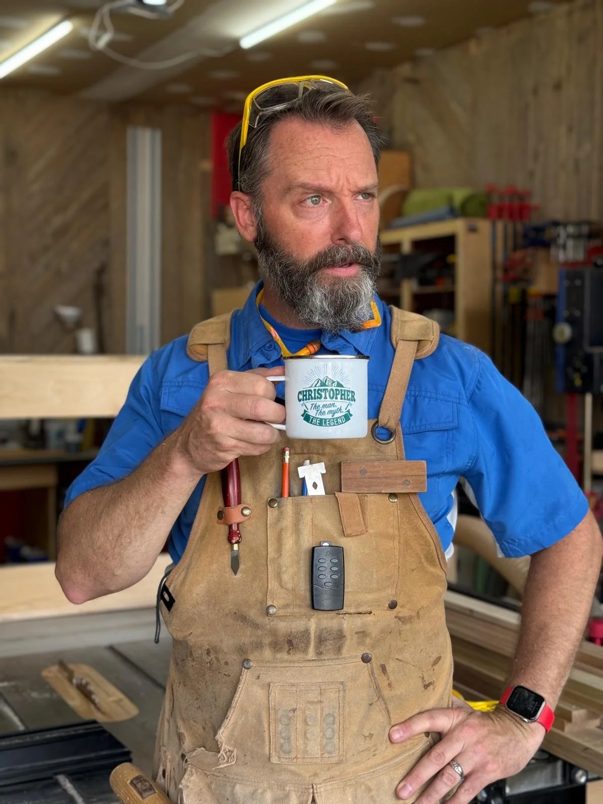 A man with a beard and glasses on his head, wearing a blue shirt and a brown work apron, stands in a woodworking shop. He is holding a mug with the label 'Christopher' and the words 'The man, The myth, The legend.'
