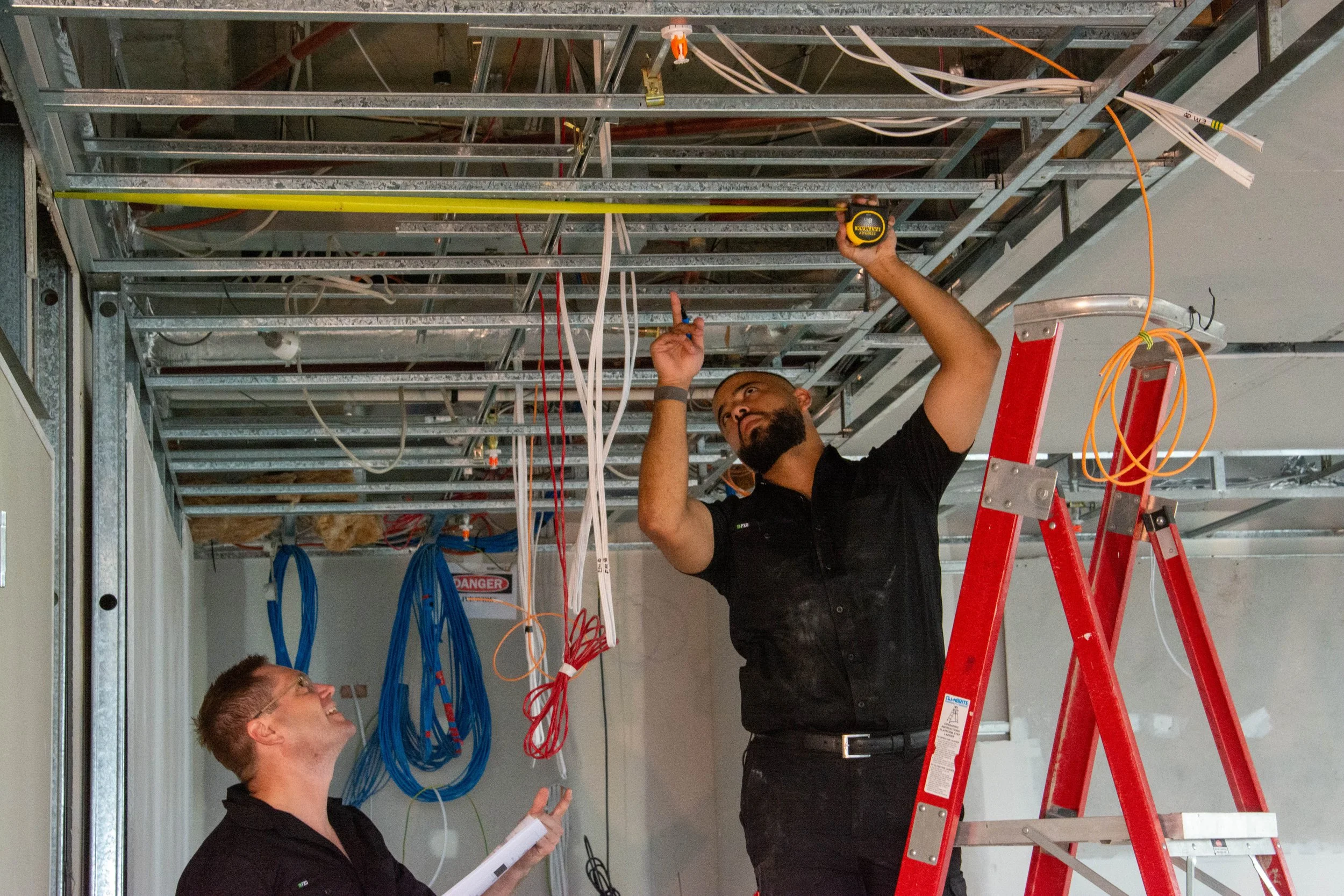 Two workers installing or inspecting electrical wiring and metal framework ceiling in an unfinished room, with one standing on a red ladder and the other looking up with a notebook.