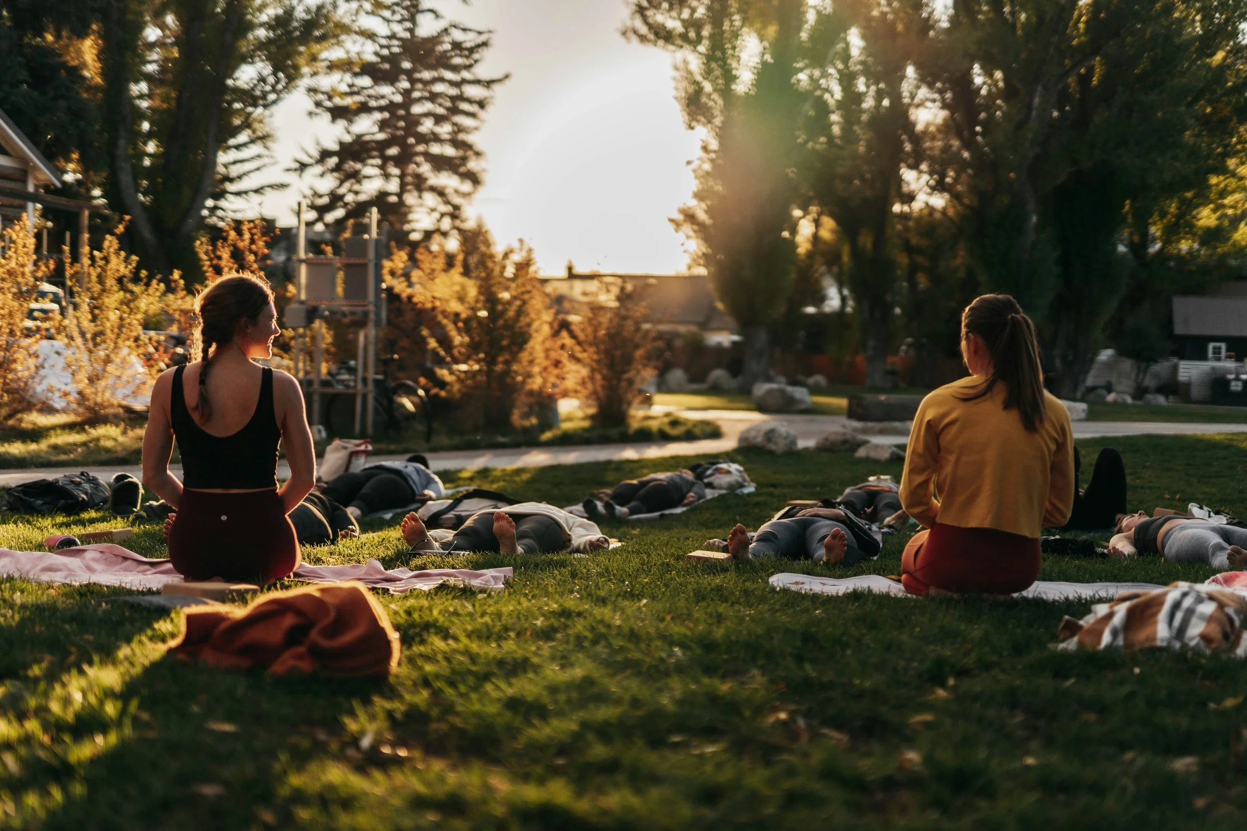 Two women are sitting on the grass during sunset in a park, participating in an outdoor meditation session with people lying on yoga mats around them, during Yoga Retreat.