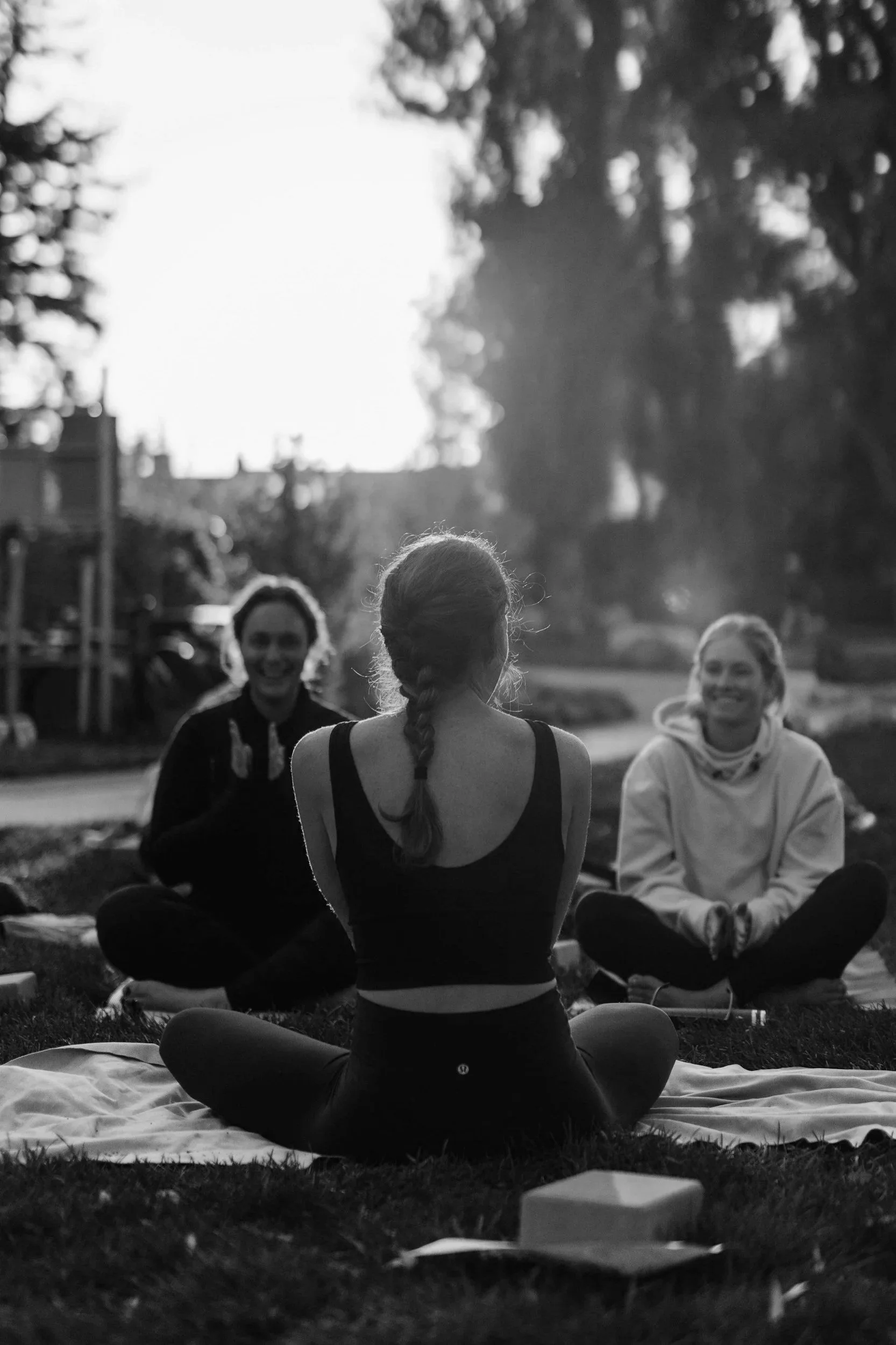 Three women practicing yoga outdoors on a grassy area in the evening, sitting cross-legged on mats and facing each other, with trees in the background, demonstrating the concept of share circles.