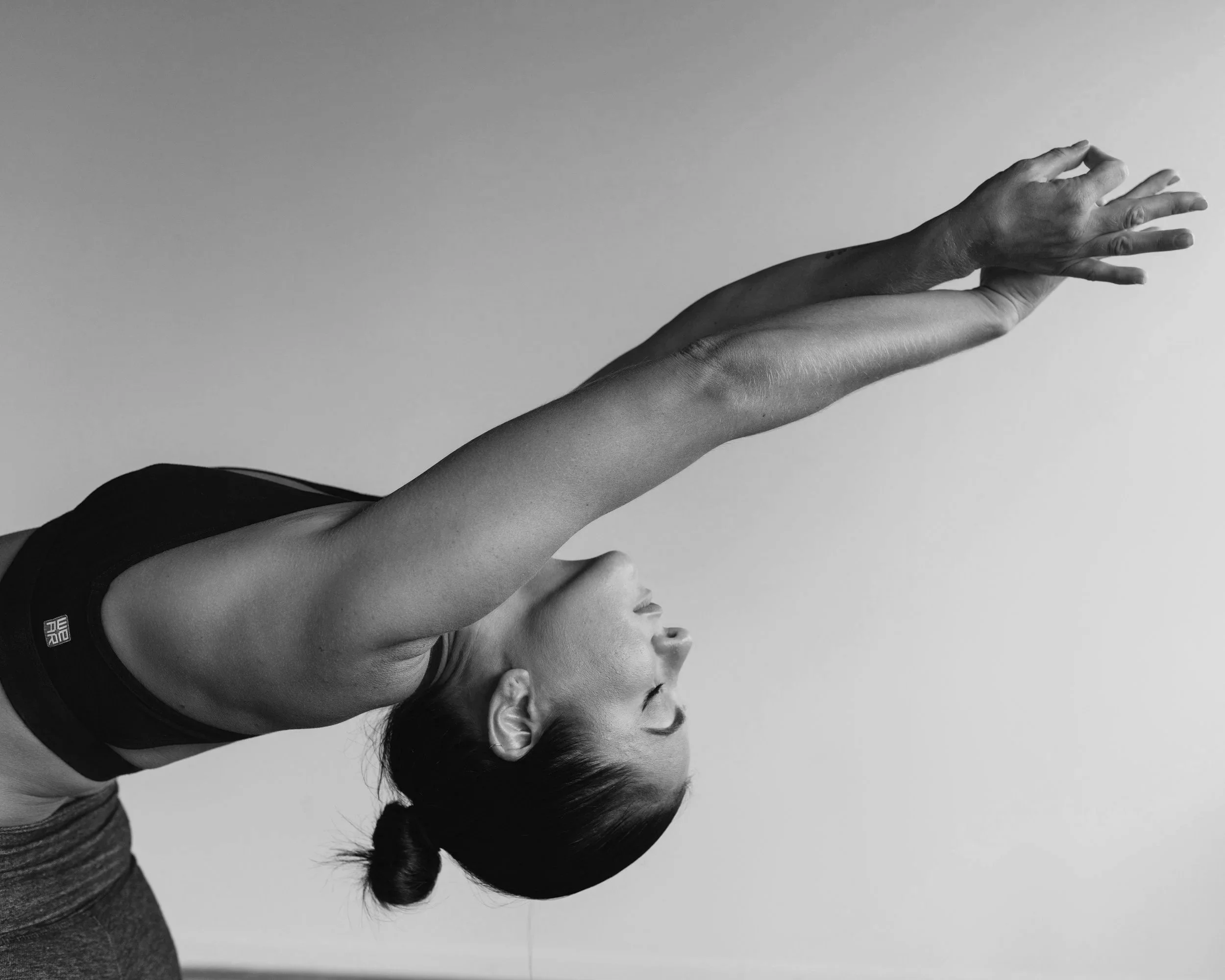 A woman in workout attire stretching with her arms reaching forward and her head tilted down, in black and white