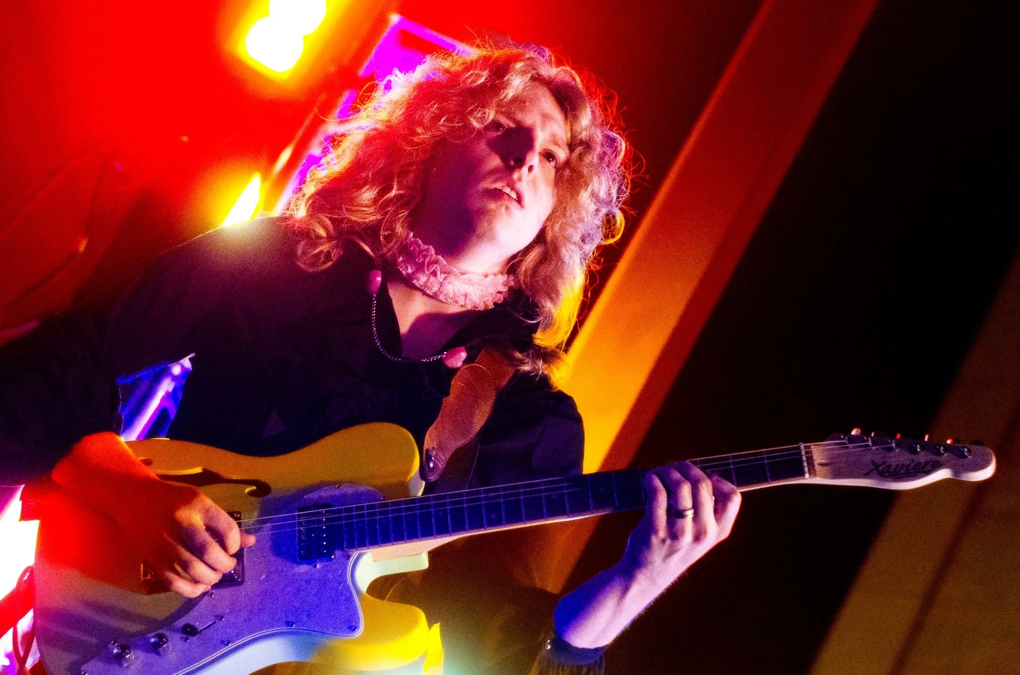 A woman with curly blonde hair playing a yellow electric guitar on stage with colorful neon lighting.