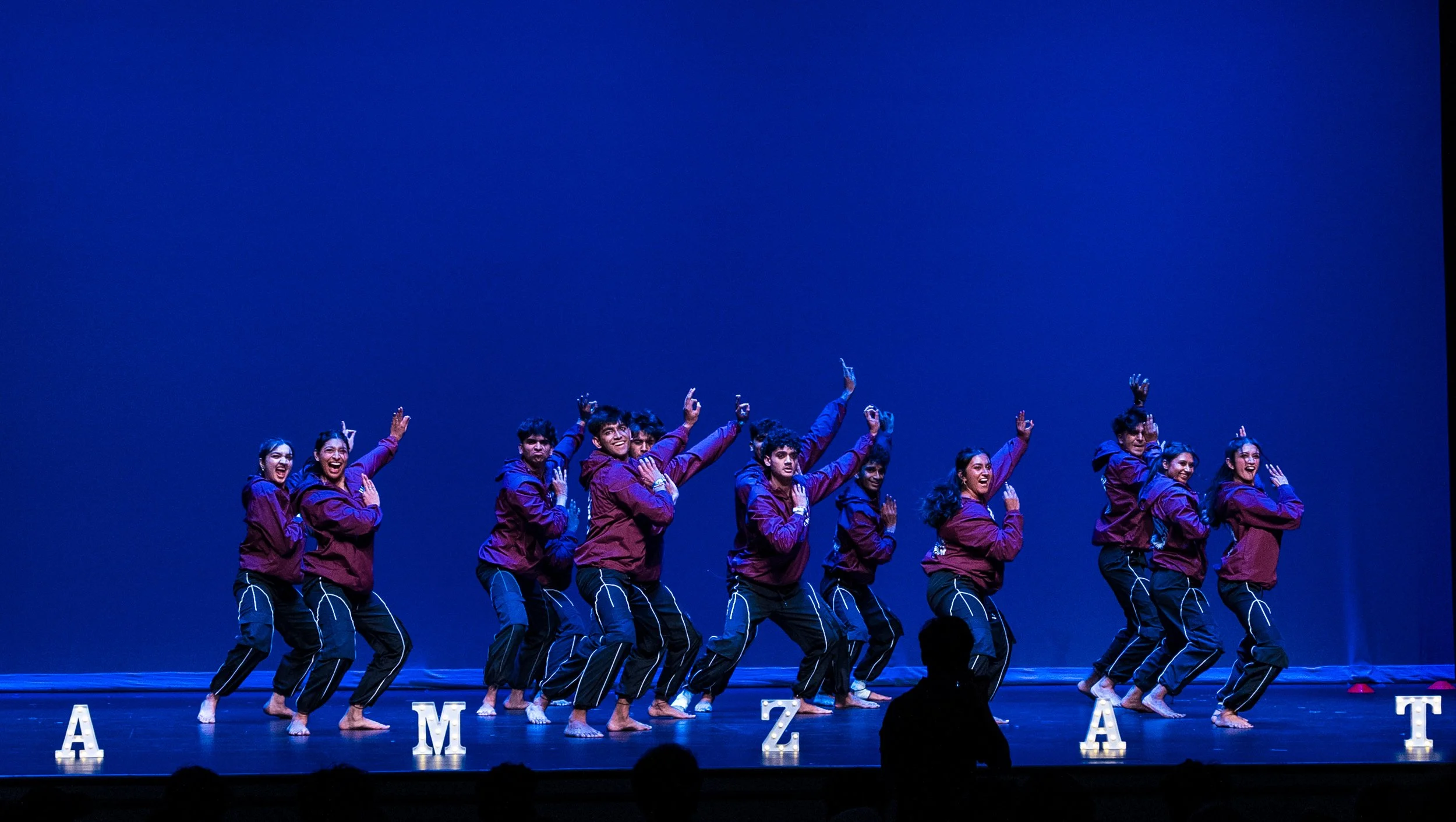 A group of dancers performing on stage with a blue background, wearing matching purple jackets and black pants with white stripe details, arranged in a line with some raising their hands and smiling.