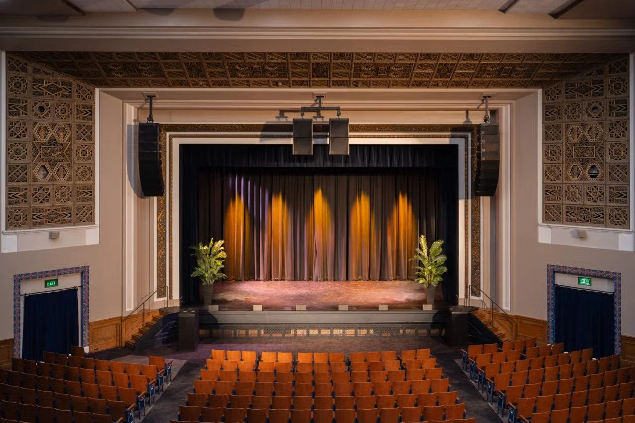 Empty theater with a stage, black curtains, potted plants, and rows of orange seats.